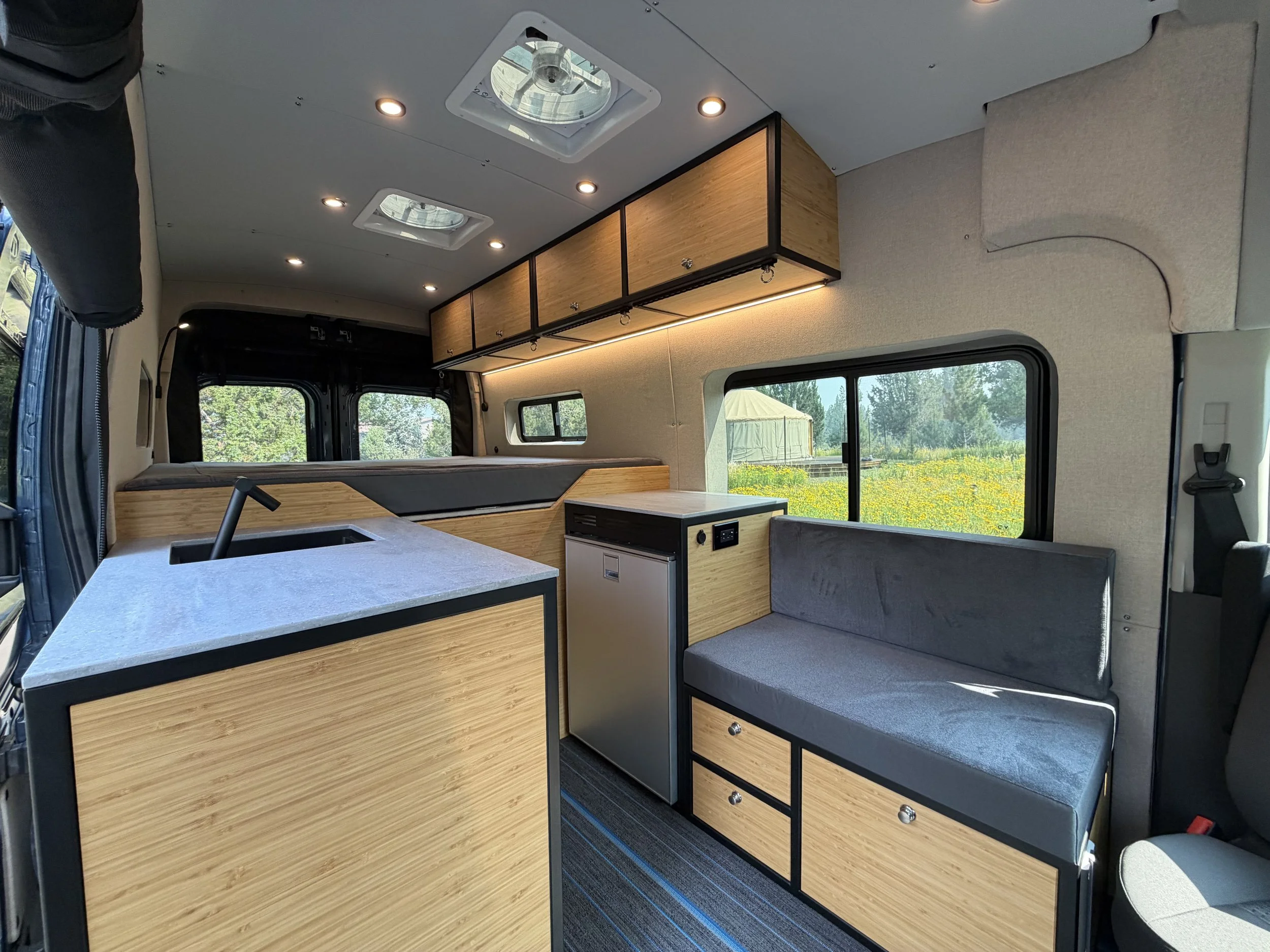 Interior of a camper van with wood cabinets, gray seating, kitchen counter, and windows showing a field and tents outside.