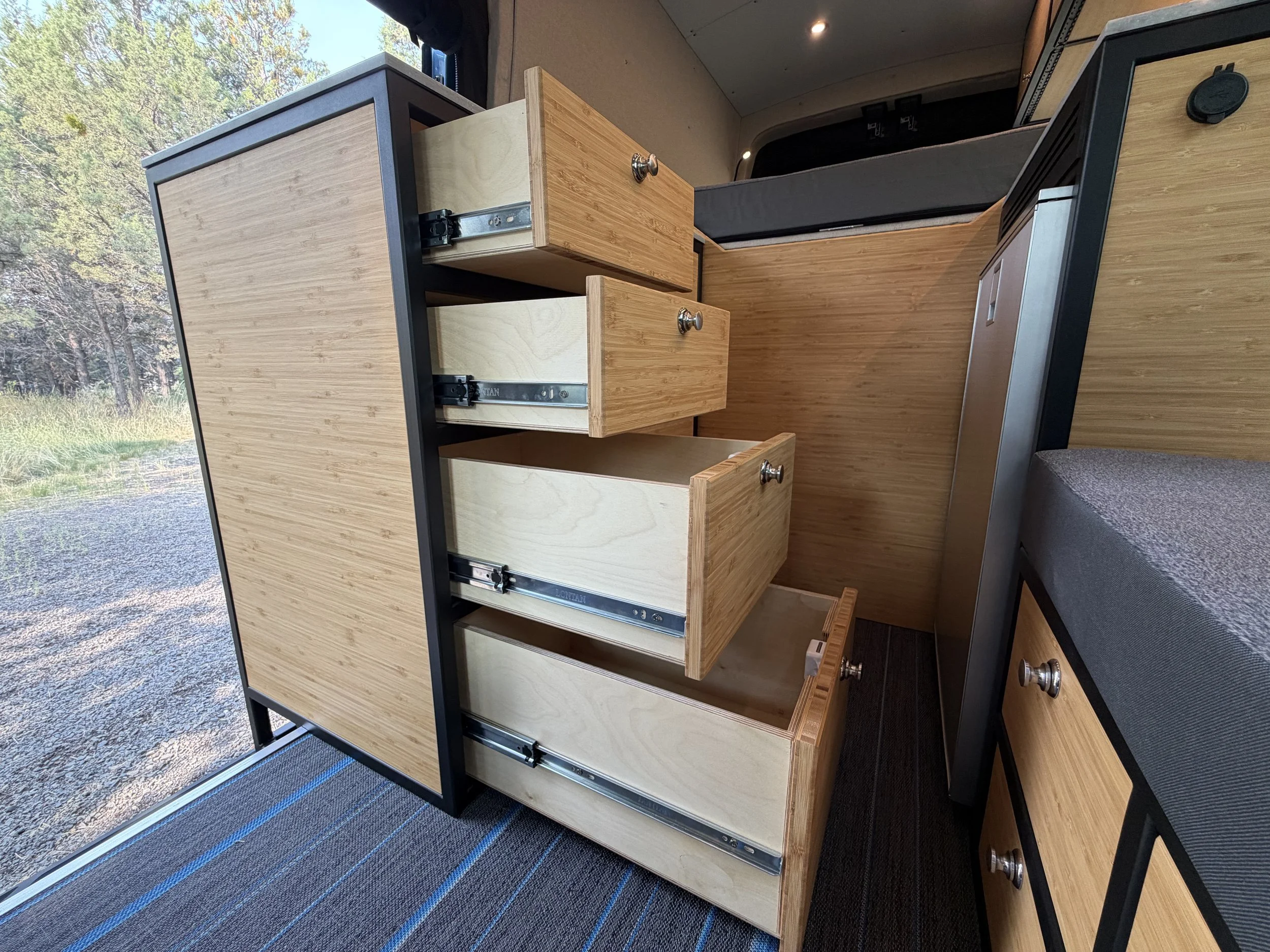 Interior of a vehicle with a wooden kitchen cabinet featuring multiple open drawers and a countertop, outdoors scene visible through the window.