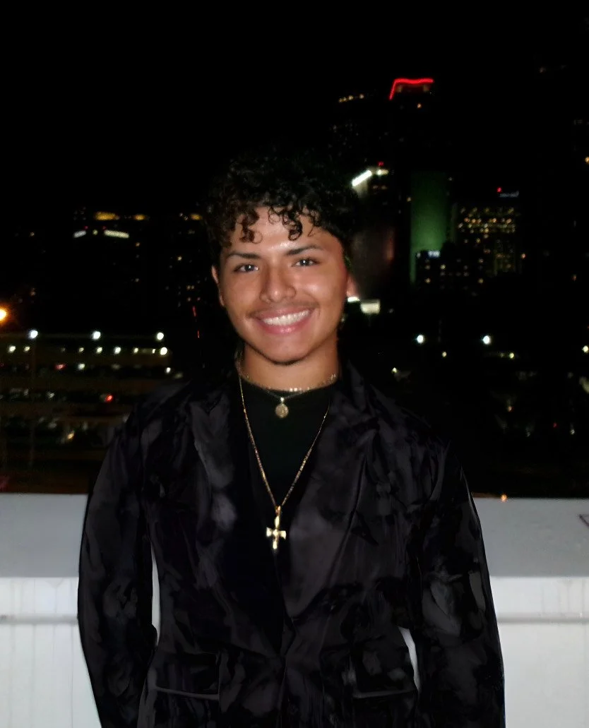 A young man with curly hair smiling at night with a city skyline in the background, wearing a black jacket and gold jewelry.