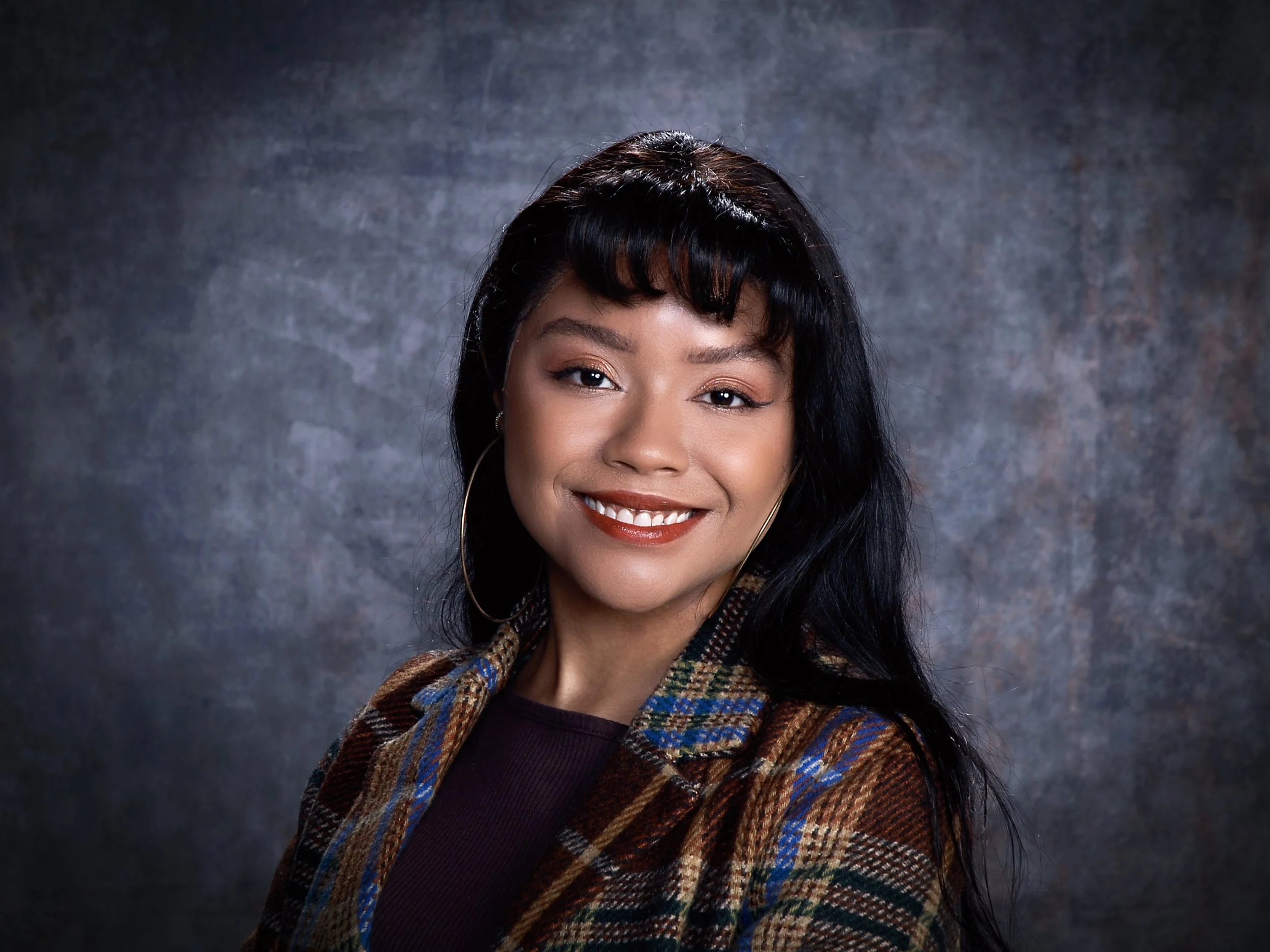 Portrait of a young woman with black hair and hoop earrings, smiling, wearing a plaid jacket, against a textured gray background.