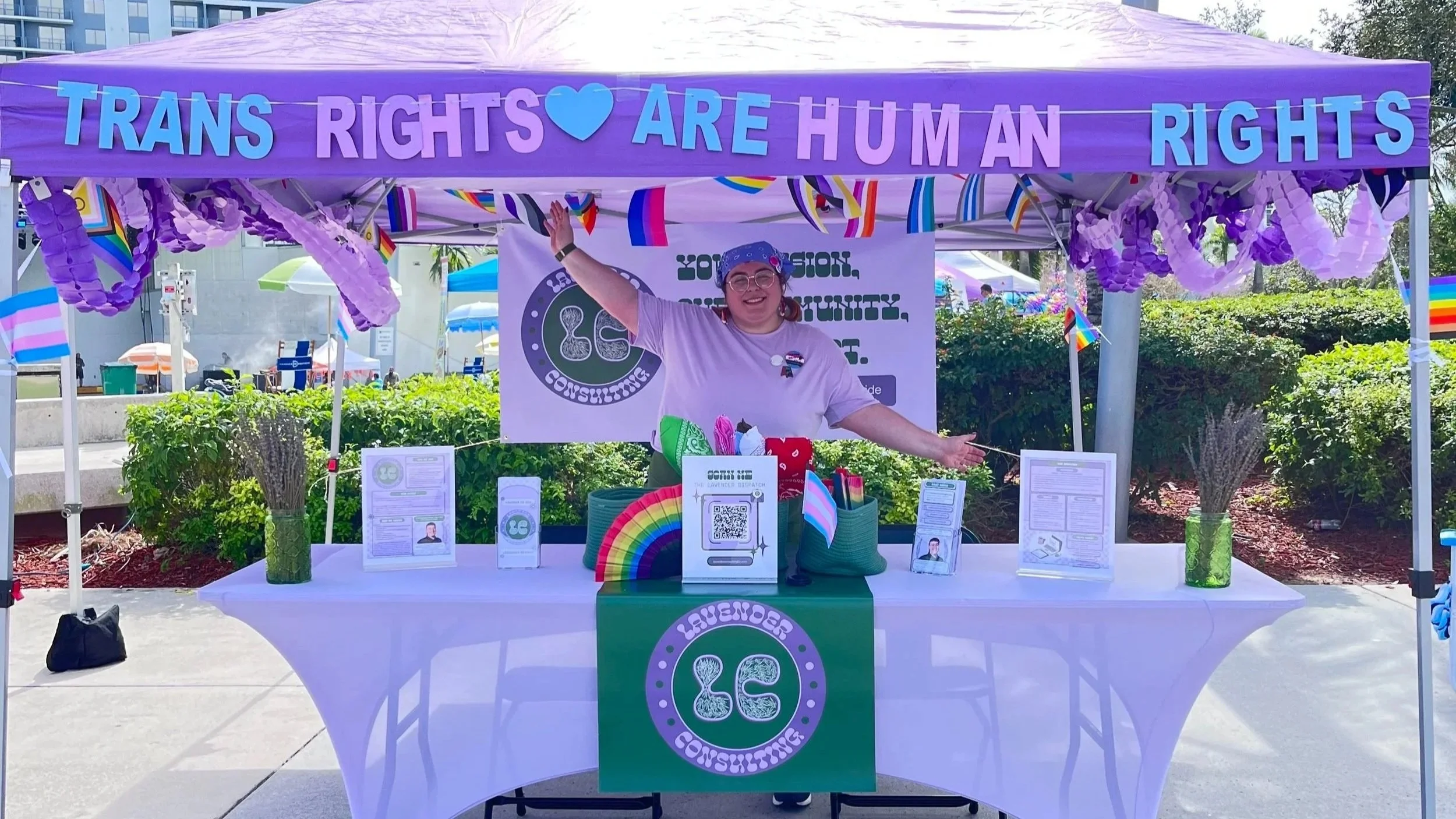A person standing behind a booth with a purple canopy that reads 'Trans Rights Are Human Rights.' The booth has various informational materials, rainbow-colored decorations, and a green banner with a logo that says 'LGB' and 'Counseling.' The person is smiling and waving, wearing a white t-shirt, glasses, and a blue bandana. The background shows outdoor setting with greenery and other booths.
