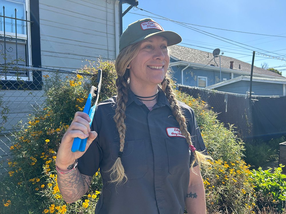 Plumber outside house wearing We Can Do It Plumbing hat and shirt and holding tools.