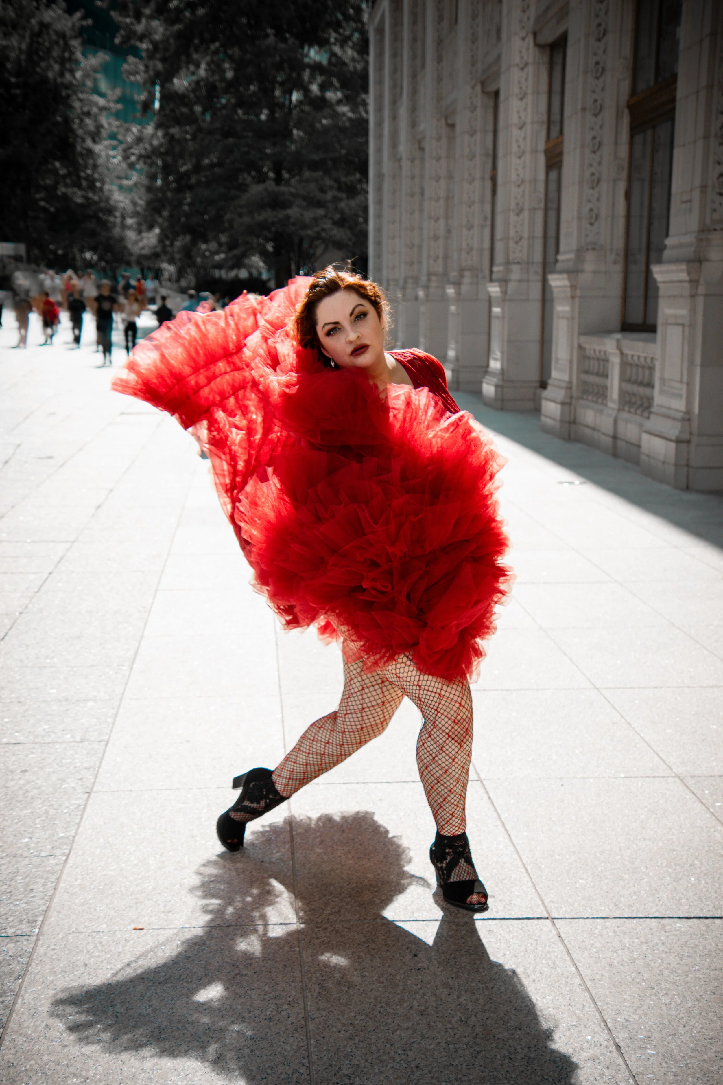 Woman in a red dress with layered tulle skirt, fishnet stockings, and high-heeled shoes poses on a city street in front of a stone building with ornate architectural details, with a group of people in the background.