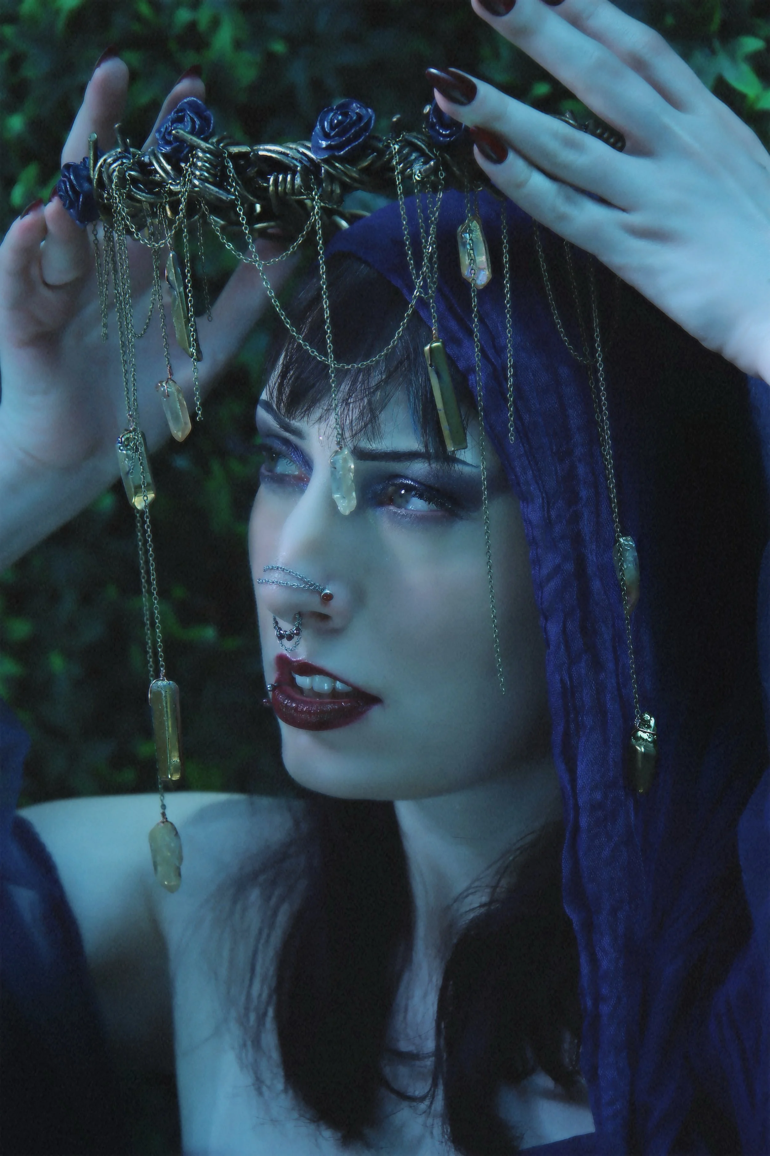 A woman with dark hair, piercings, and makeup holding a decorative chain crown with crystals and gold chains above her head, outdoors in a dark setting with greenery in the background.