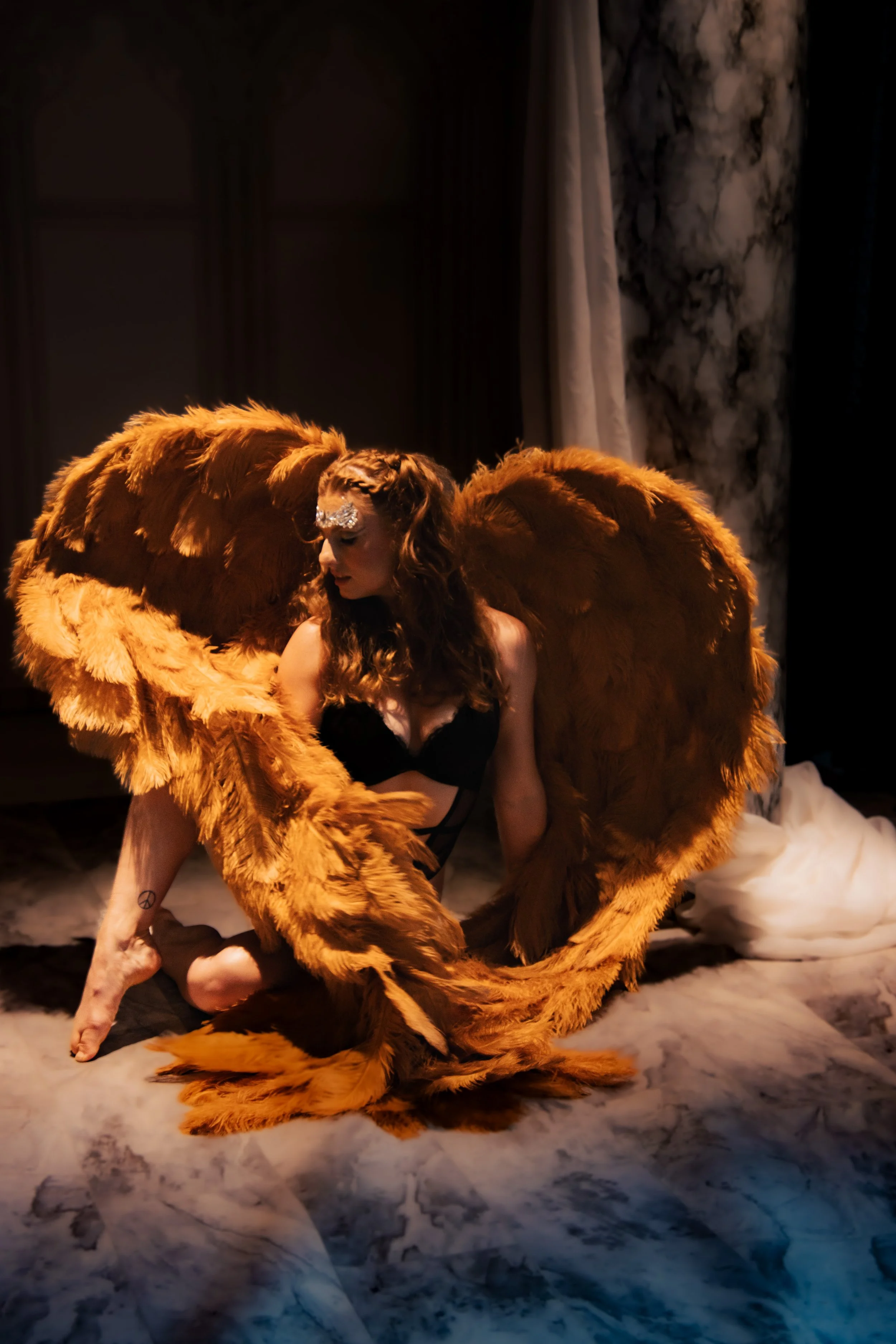 A woman with long curly hair, dressed in black lingerie and makeup, is kneeling on the floor with large brown feathered angel wings attached to her back. The background features dark, marble-like curtains and draped fabric.