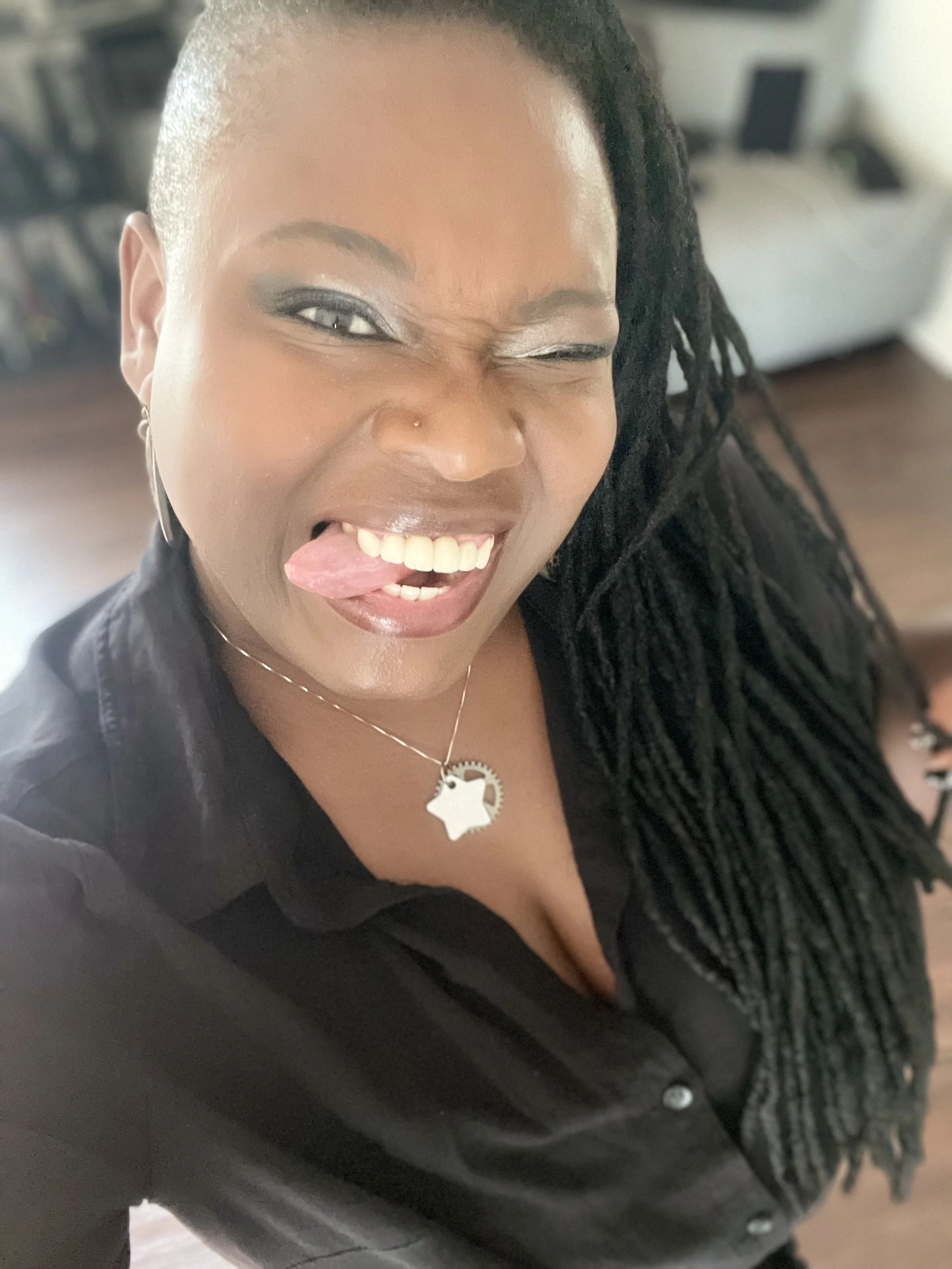 A woman with long locs and wearing a black shirt, making a funny face by sticking out her tongue and winking, wearing a gear and star-shaped pendant engraved with the name Kinesis.