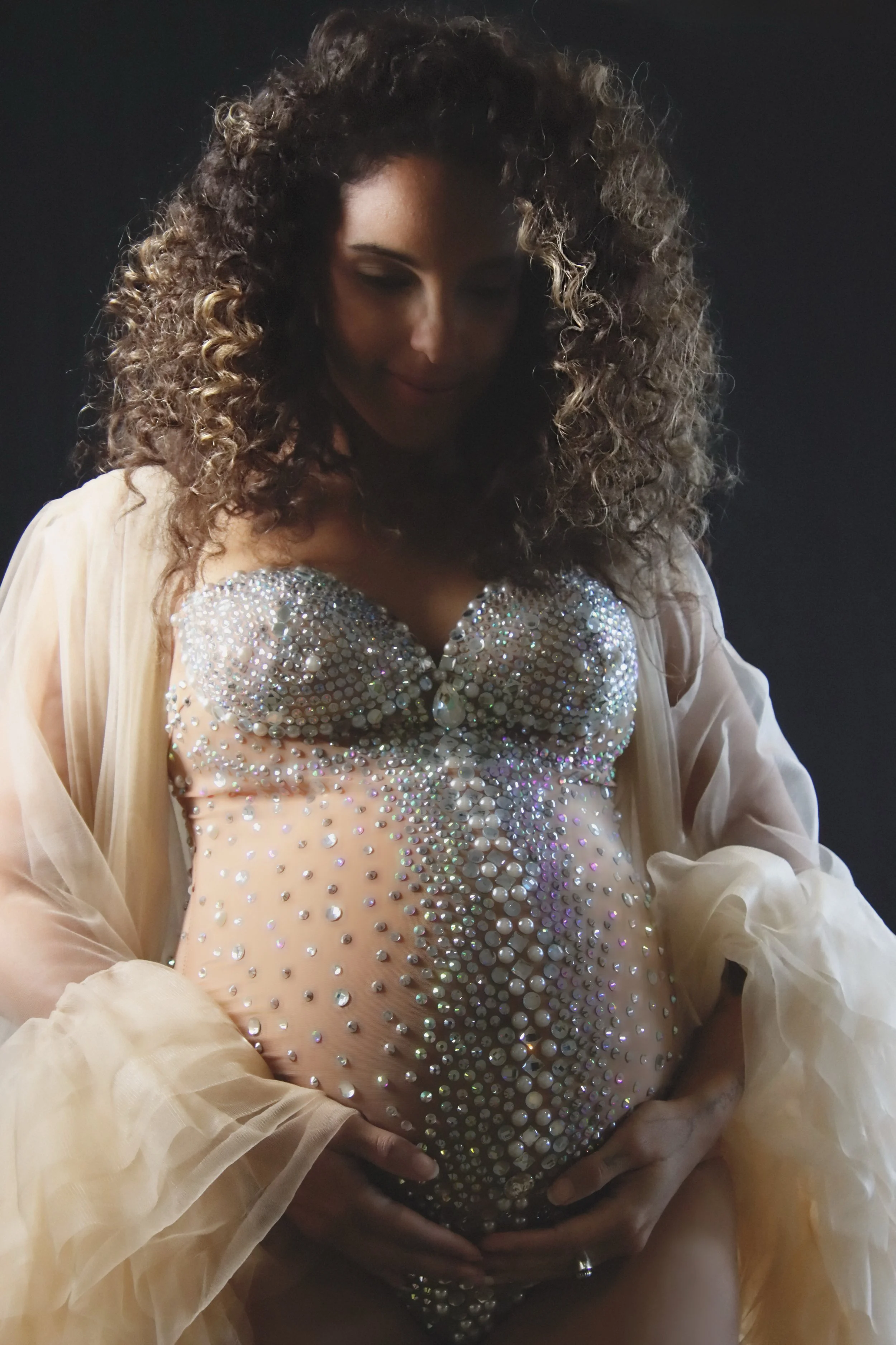 A pregnant woman with curly hair wearing a rhinestone decorated top and a sheer, beige robe, holding her belly with both hands and looking down.