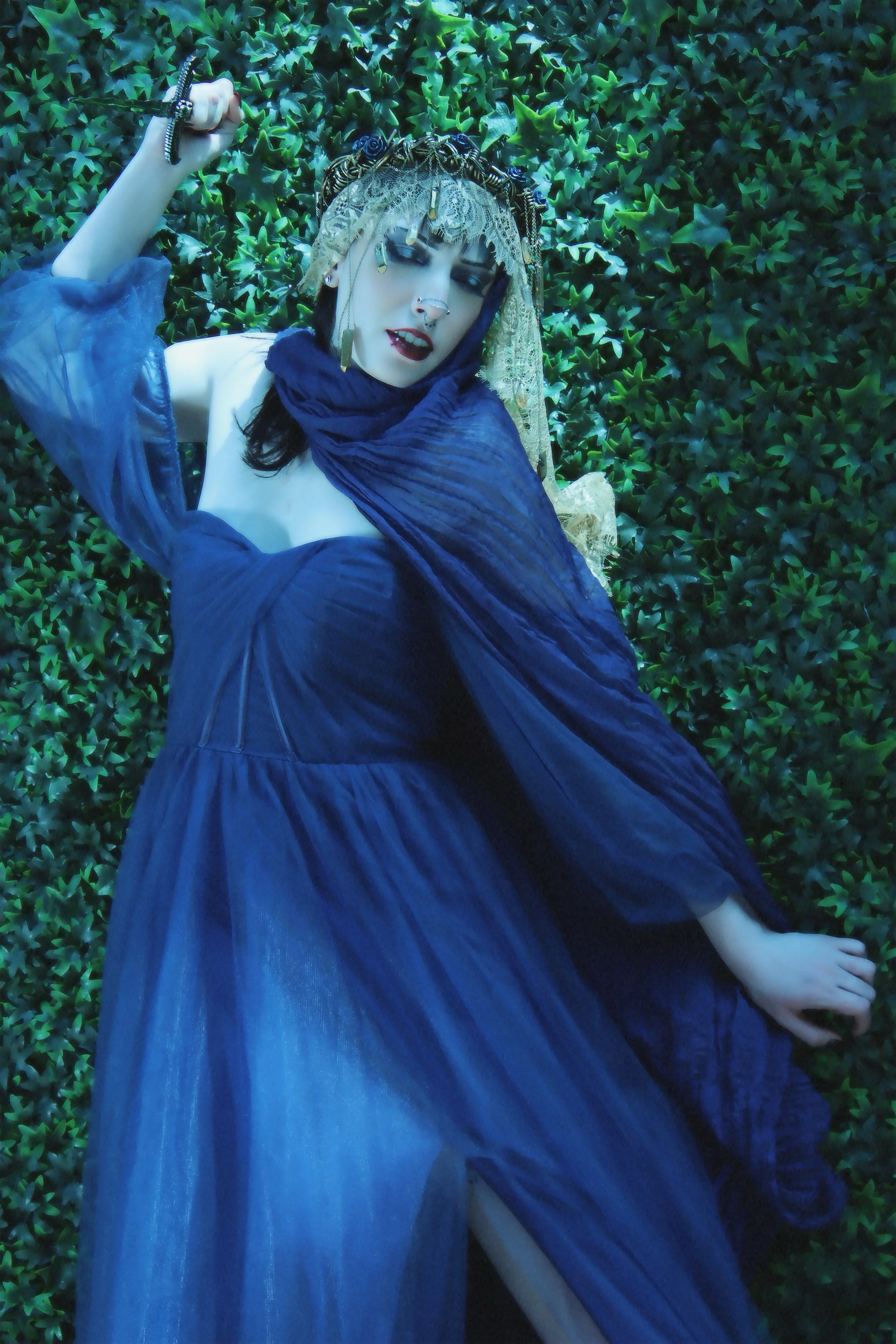A woman lying on a leafy green background, dressed in a blue gown with sheer long sleeves, wearing a veil and ornate jewelry, with makeup and jewelry, holding a dagger.