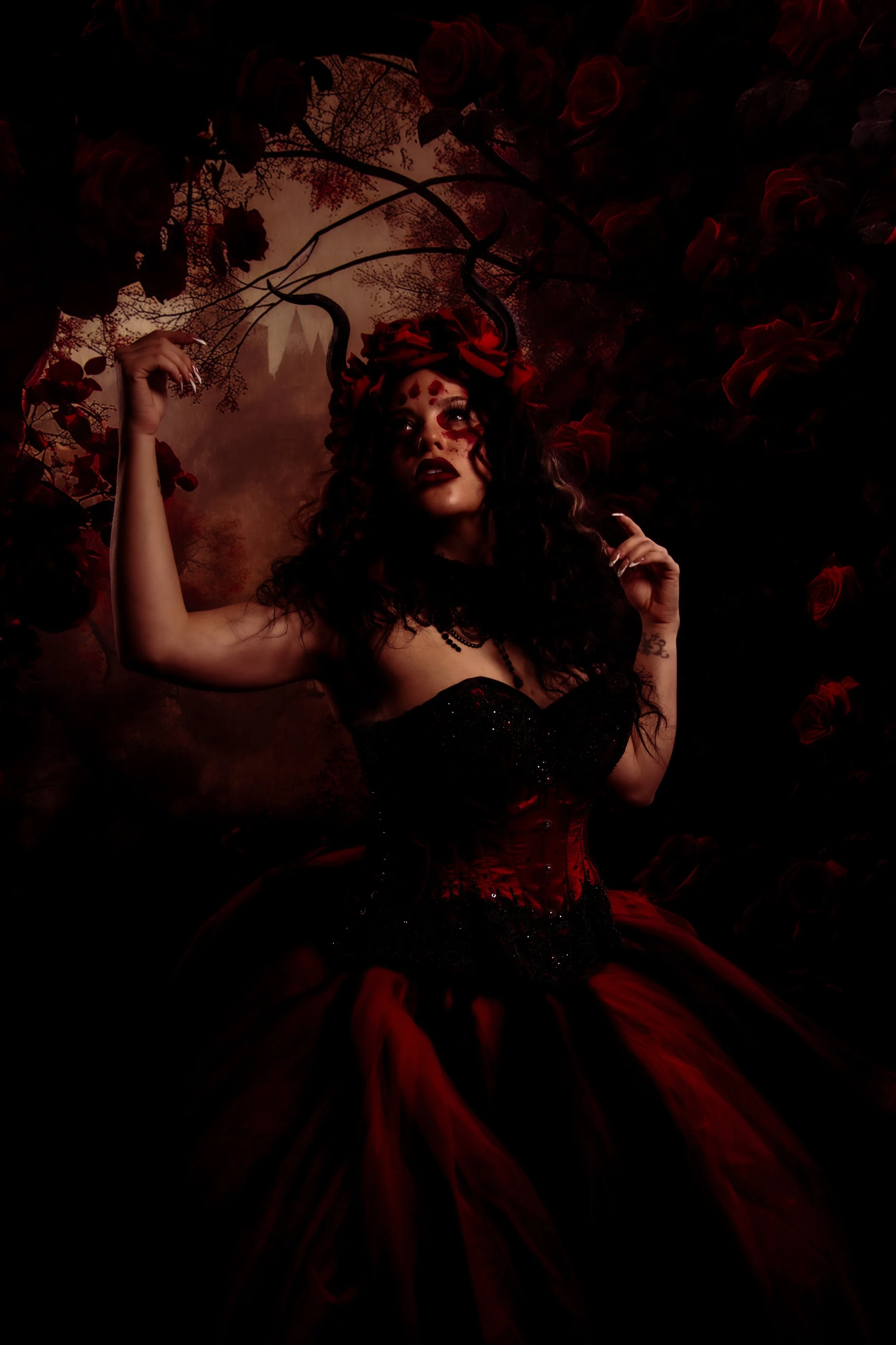 A woman dressed as a dark fairy or demon queen in a black and red corset and tulle skirt, with dark curly hair, horns, and red makeup, surrounded by roses and dark foliage.