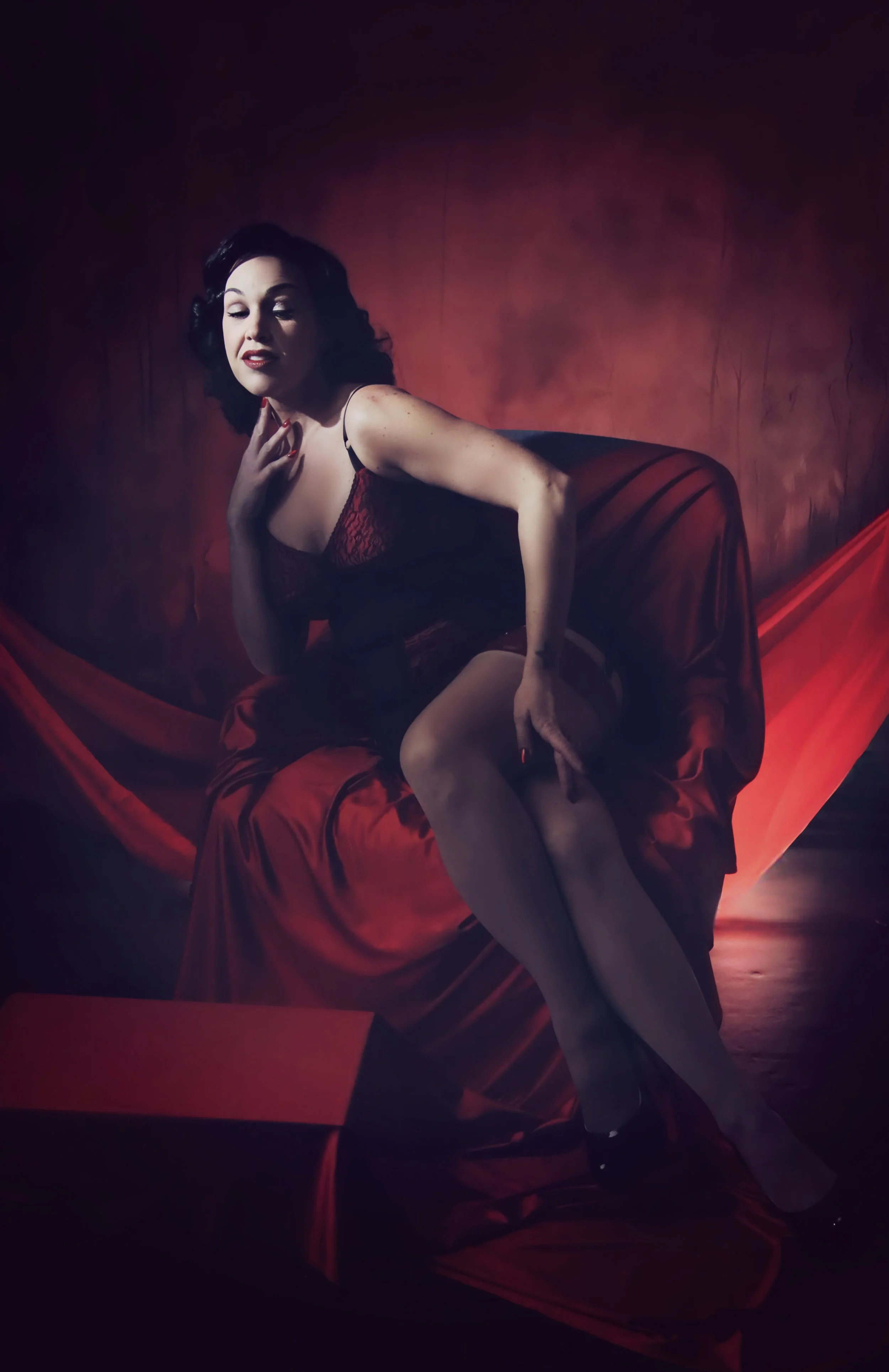 A woman dressed in vintage style lingerie with dark hair, sitting on red satin fabric against a dark, moody background. She is posed sensually with her legs crossed and her hand near her face.