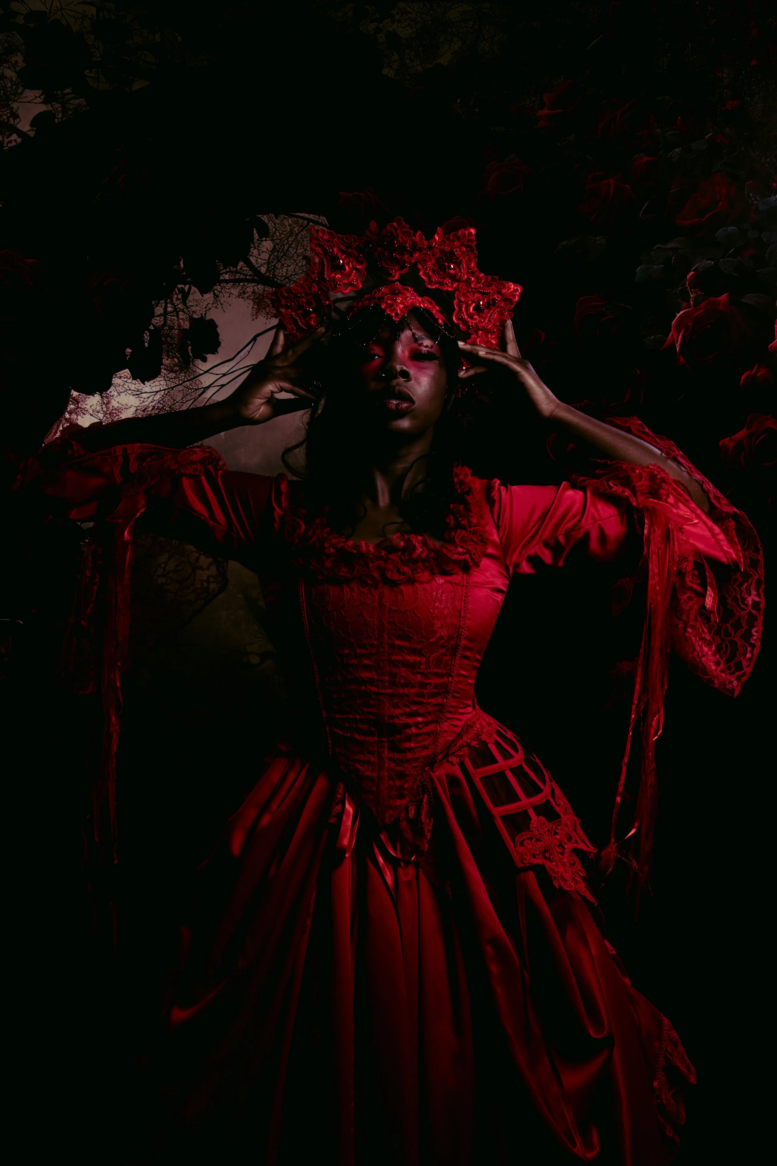 A woman in a red dress and Gothic-style costume lying on the ground among roses and dark foliage, holding her head with her hands.