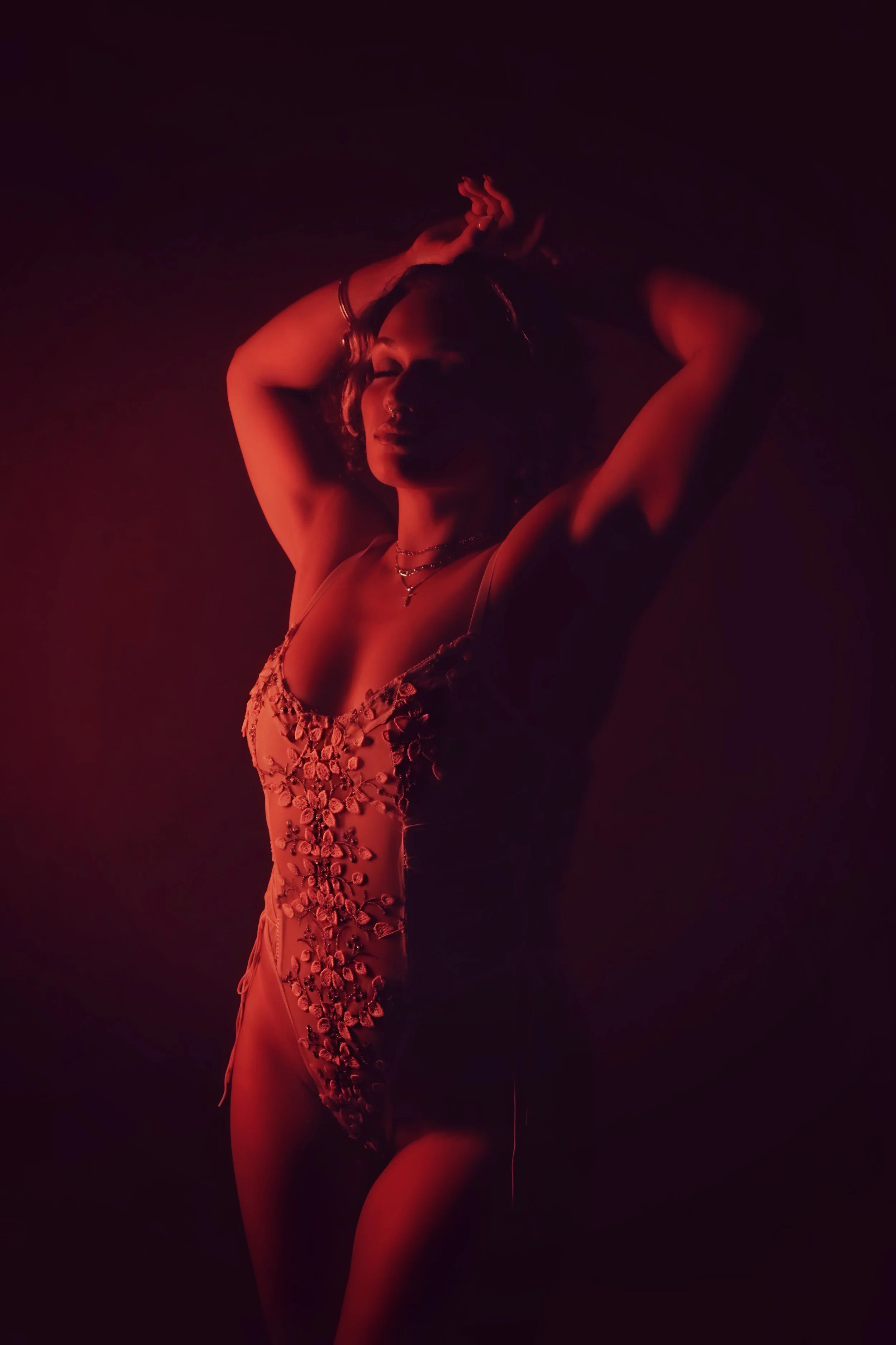 A woman with short hair wearing a floral embellished dress and jewelry, posing with arms raised against a dark red backdrop, illuminated by red lighting.