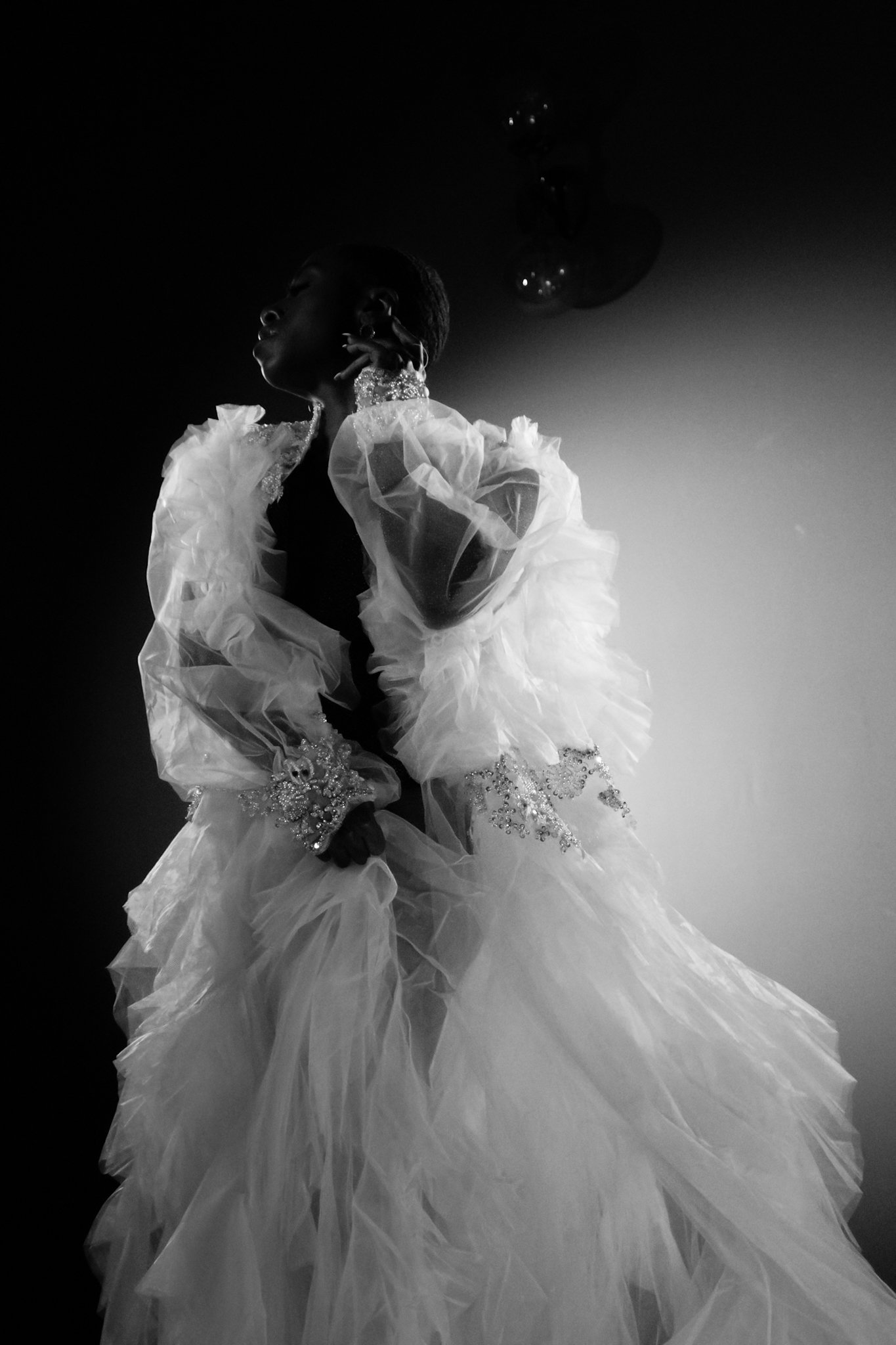 A black and white photo of a woman in an elaborate, ruffled dress, holding the skirt with one hand, with the other hand near her neck, gazing upward with a serious expression.