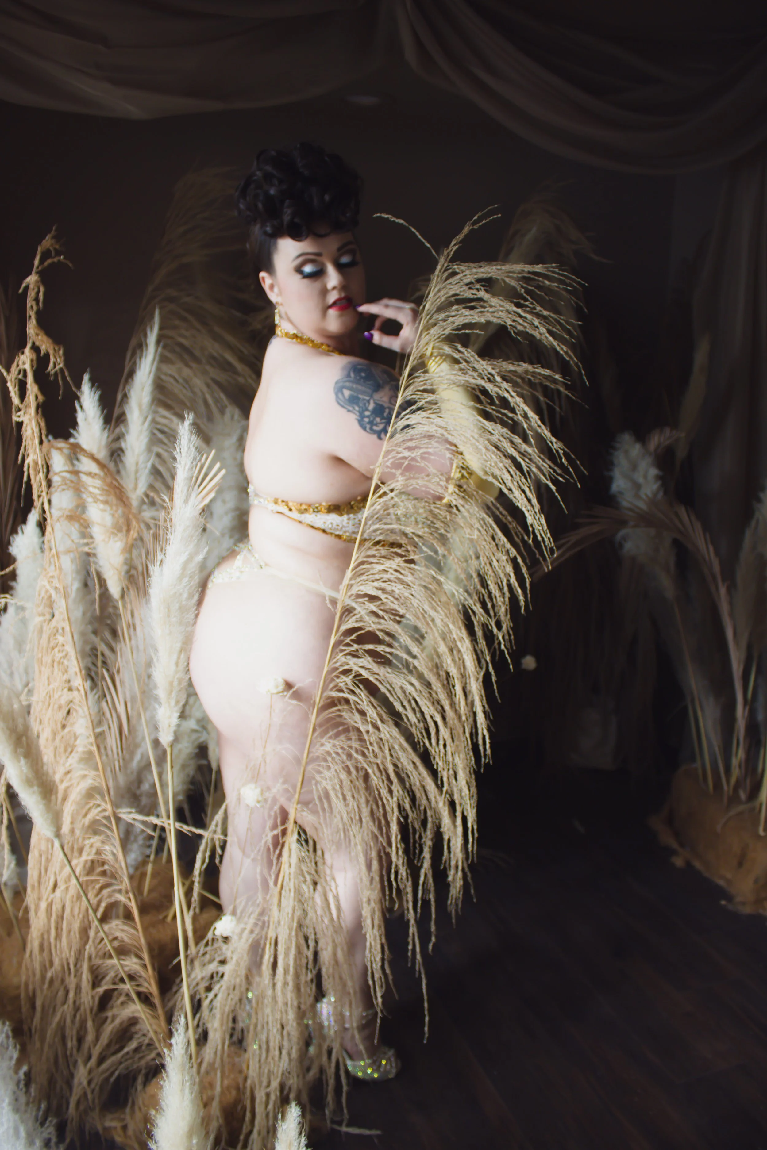 A woman with dark curly hair, tropical makeup, and tattoos, standing among tall beige pampas grass, wearing gold jewelry and glittery high heels, with a dark background and draped fabric overhead.