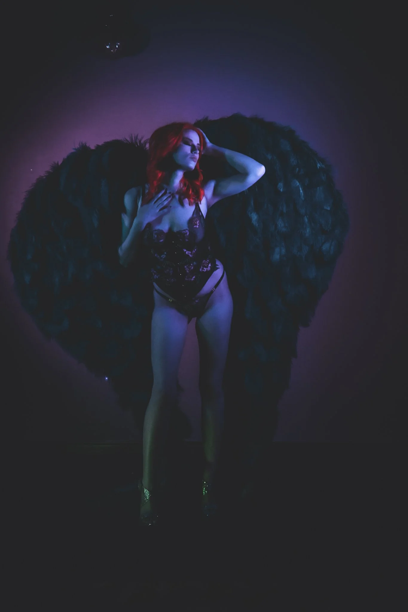 A woman with red hair poses against a purple background with black wings behind her, wearing black lingerie and gold high heels.