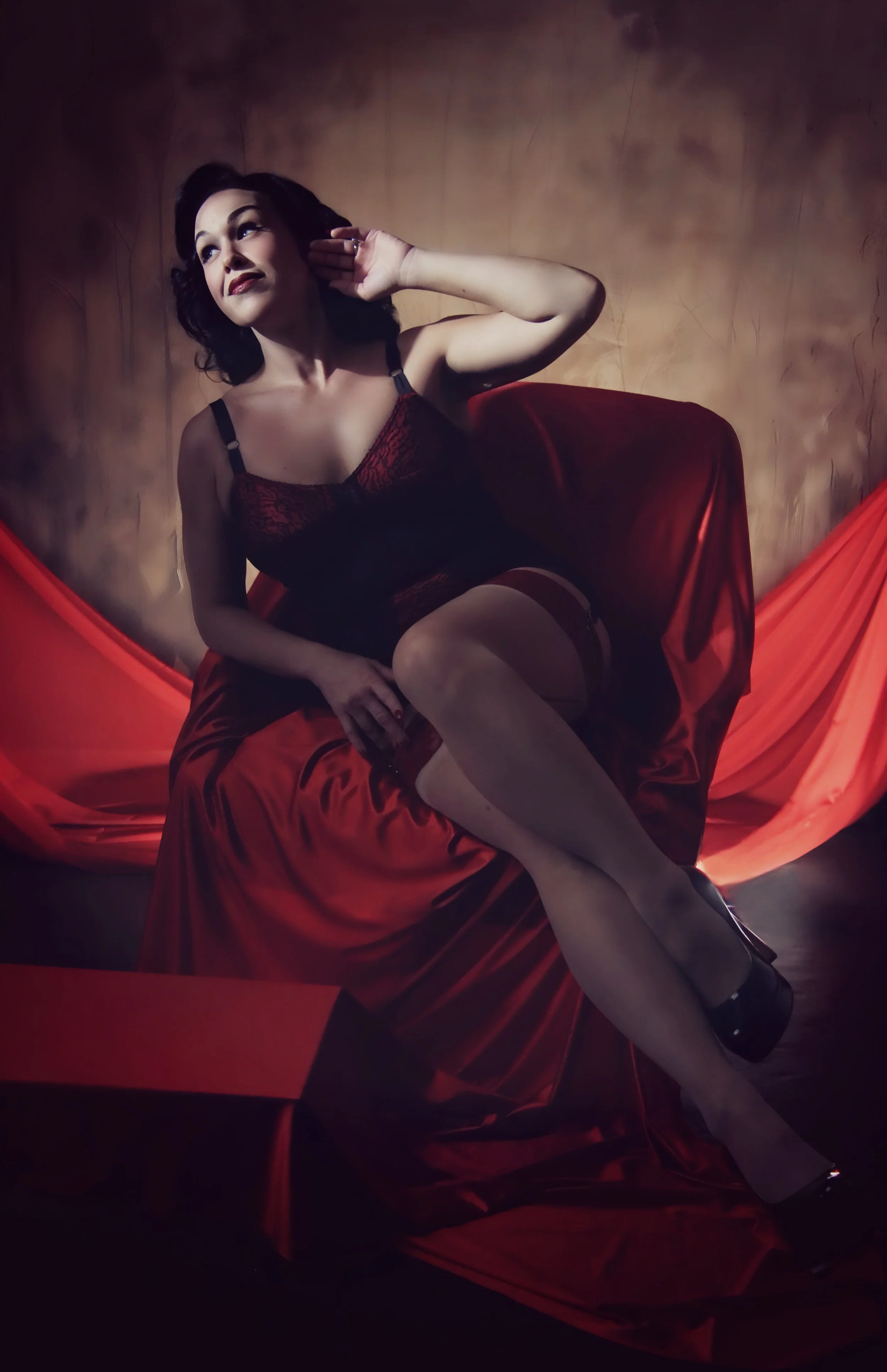 A woman with dark, wavy hair wearing black and red lingerie, sitting on a red satin-covered chair with a red drape behind her, against a textured brown backdrop.