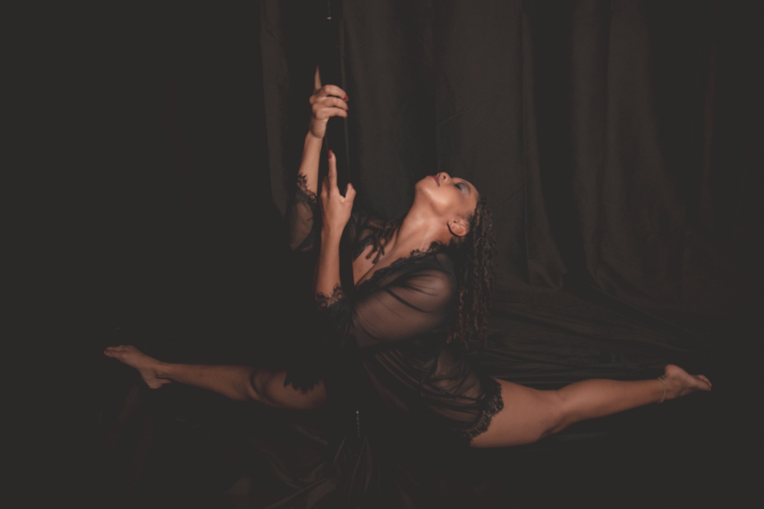 A woman with curly hair in black sheer lace dress performing a split on dark fabric with a curtain backdrop.