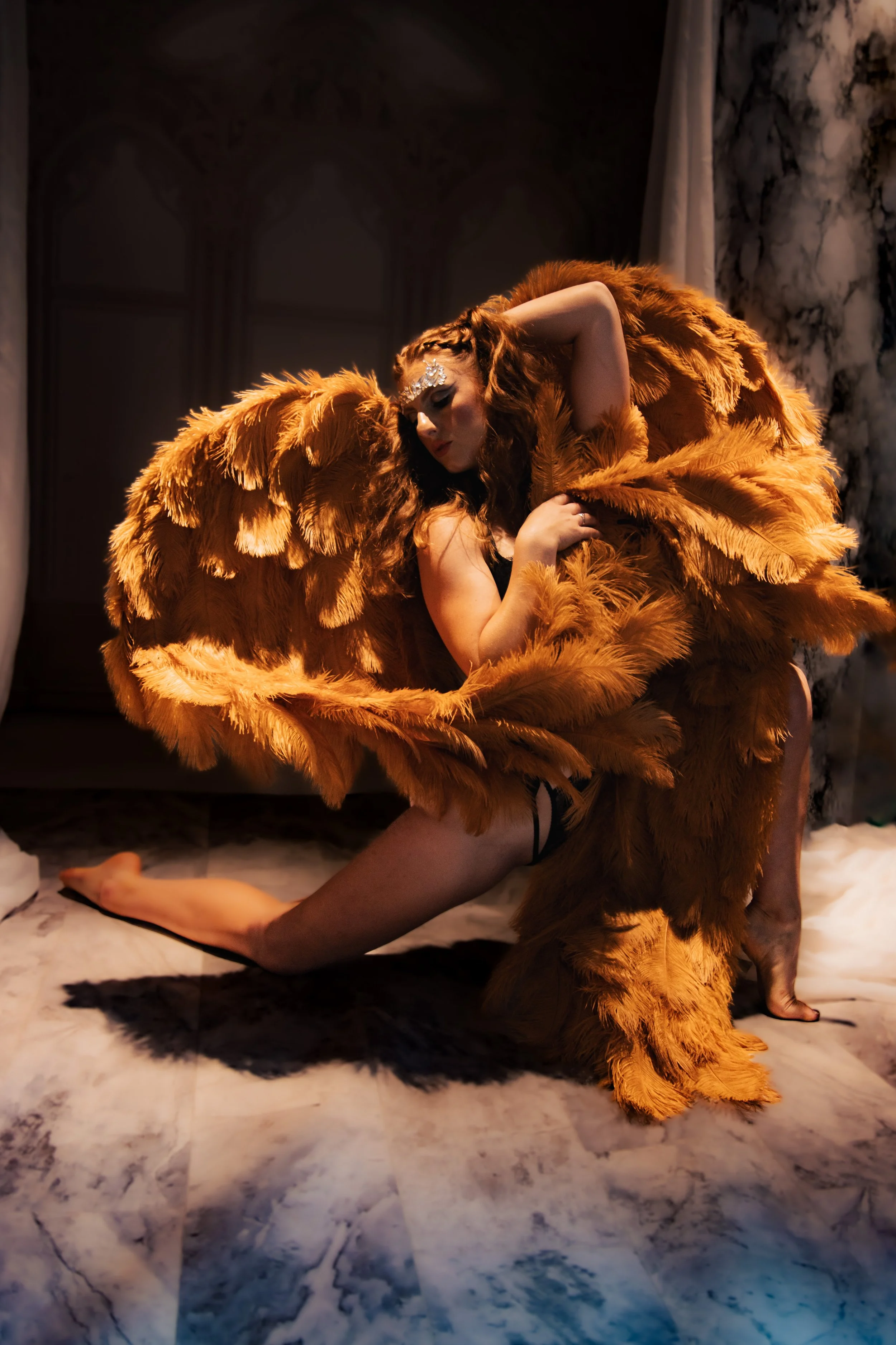 A woman with curly red hair in a black outfit and feathered orange costume, posing on a marble floor with a dark background and marble wall, with dramatic lighting.