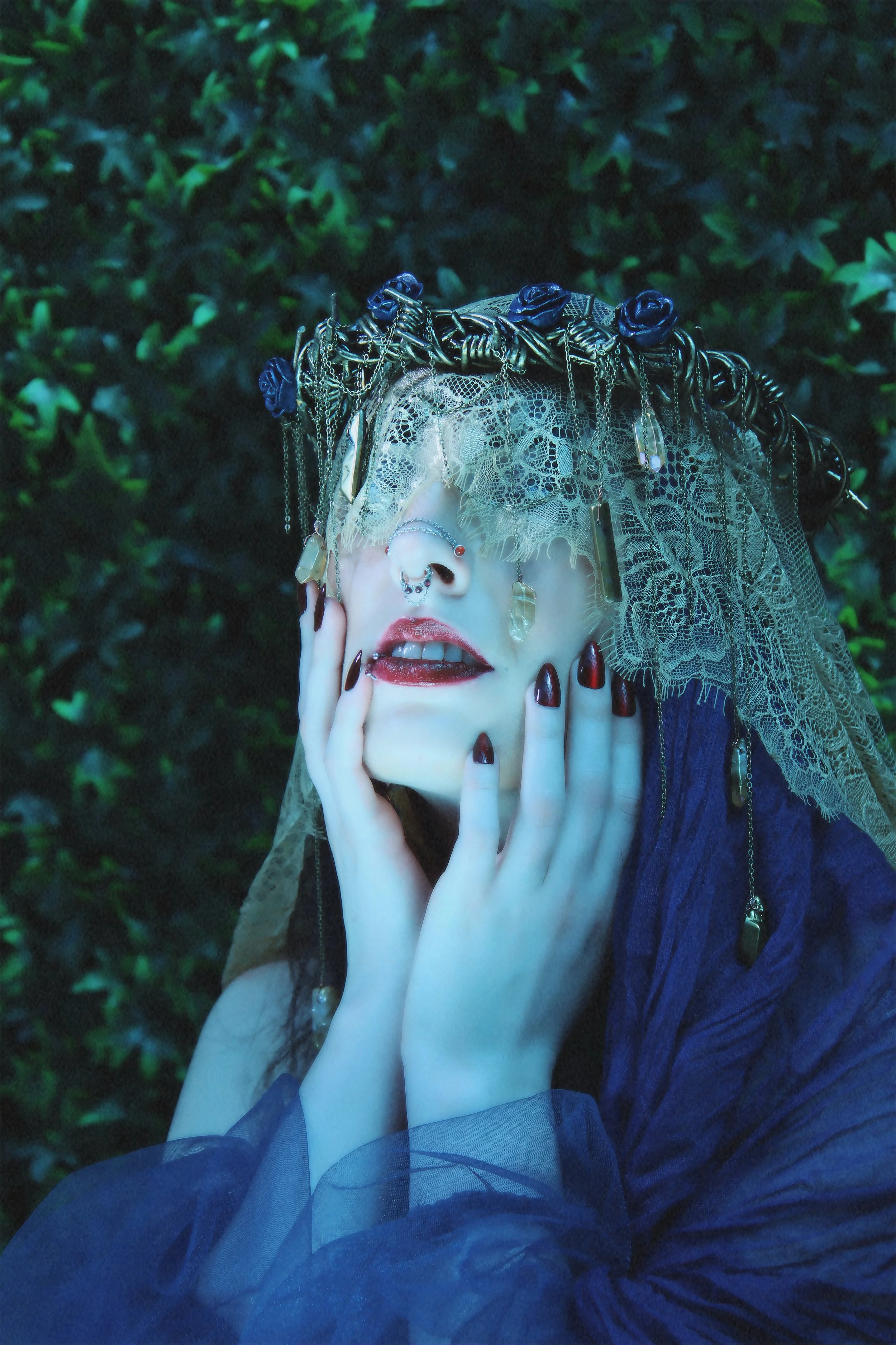 A woman with pale skin, dark lipstick, and dark eyeshadow, wears a lace veil and intricate jewelry, including chains, crystals, and a nose ring. She touches her face gently with both hands, and her background features dark green leafy bushes.