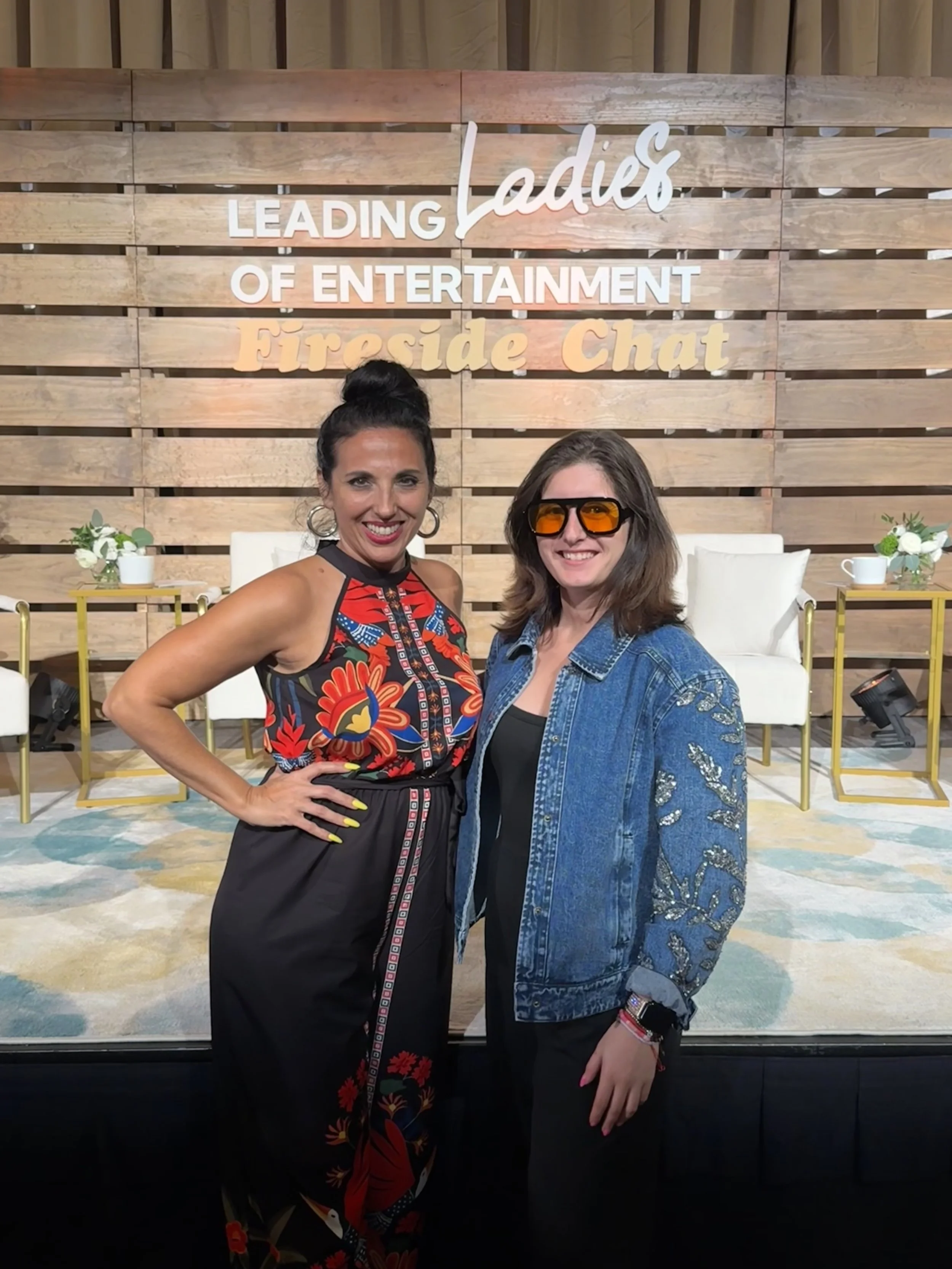 Two women smiling and posing in front of a sign that reads 'Leading Ladies of Entertainment Fireside Chat' on a wooden-paneled background.