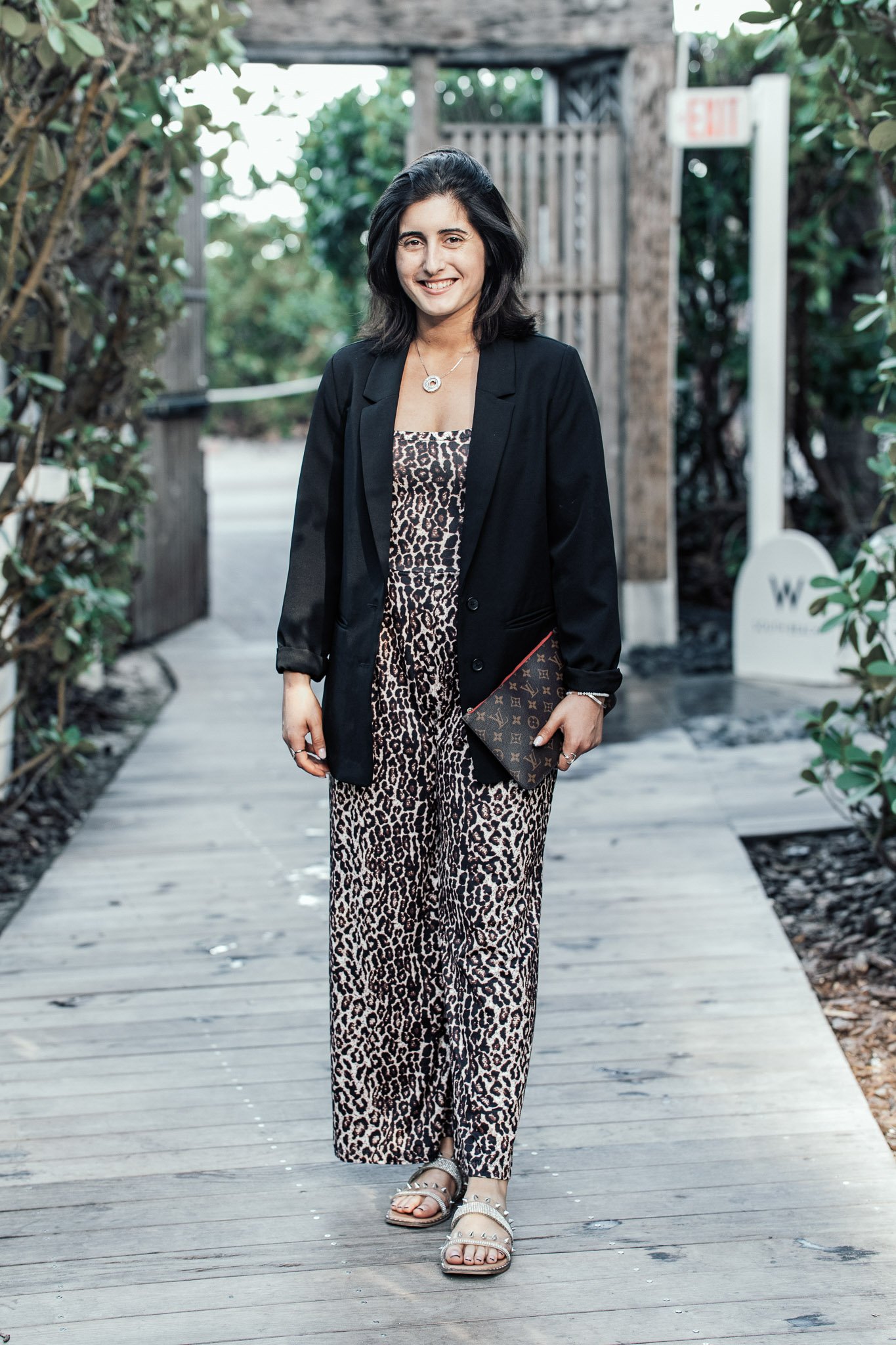 A woman with dark shoulder-length hair smiling, dressed in a black blazer over a leopard print dress, holding a Louis Vuitton clutch, wearing sandals, standing on a wooden pathway outdoors with greenery in the background.