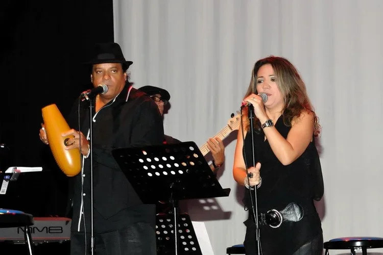 A man and a woman performing music on stage. The man is playing a percussion instrument and singing into a microphone, wearing a black hat and shirt. The woman is singing into a microphone, dressed in black with a belt, and has long, wavy hair. There is a music stand and musical equipment on stage with a white curtain background.