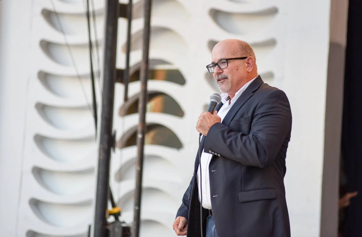 A middle-aged man with glasses and a mustache, dressed in a dark blazer and white shirt, is holding a microphone and speaking at an event. Behind him is a white wall with decorative cutouts.