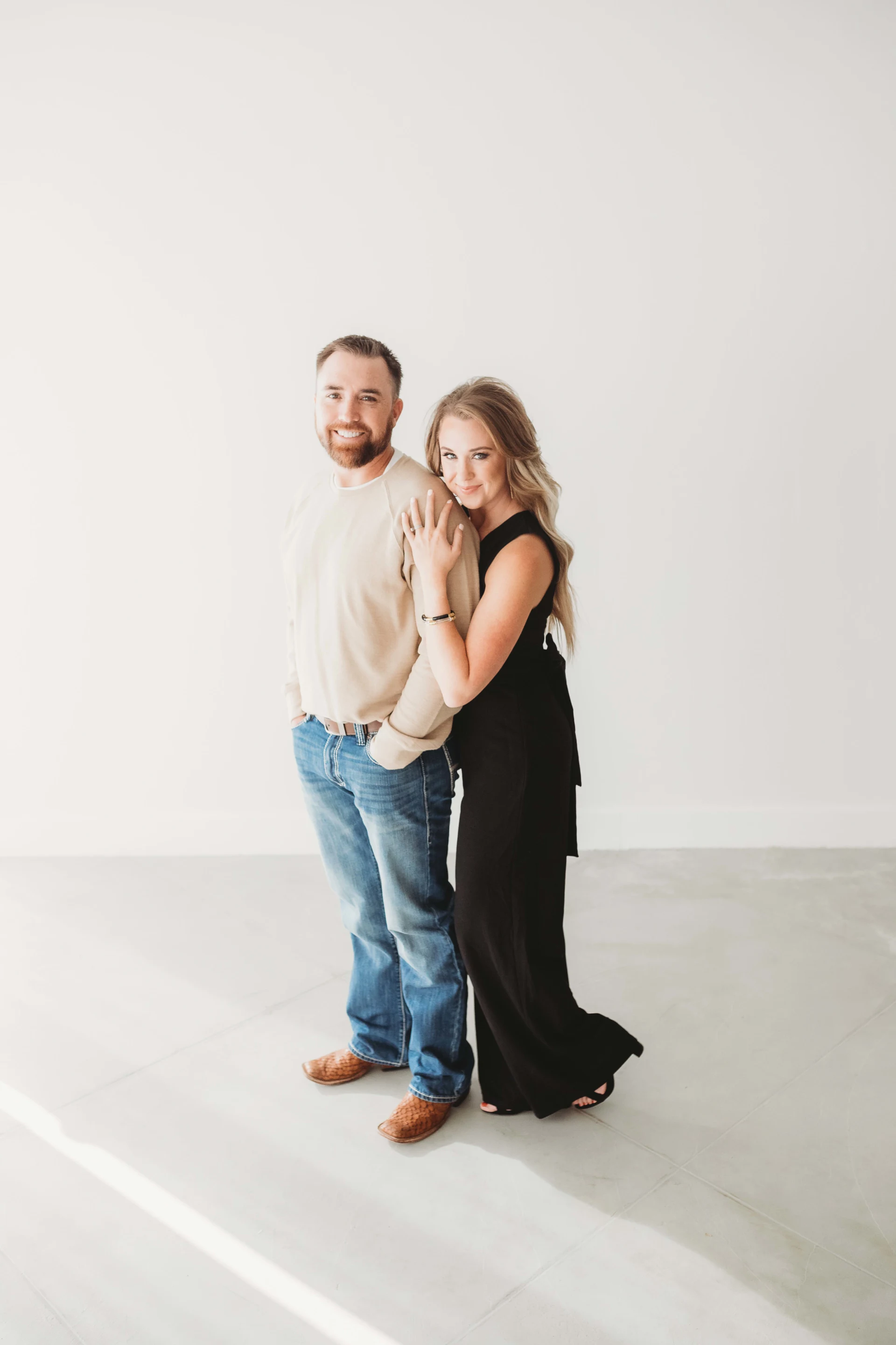 A couple posing together in a bright, minimalist room. The man is wearing a beige sweater, blue jeans, and brown shoes, smiling with hands in his pockets. The woman is leaning on him, dressed in a long black dress, also smiling.