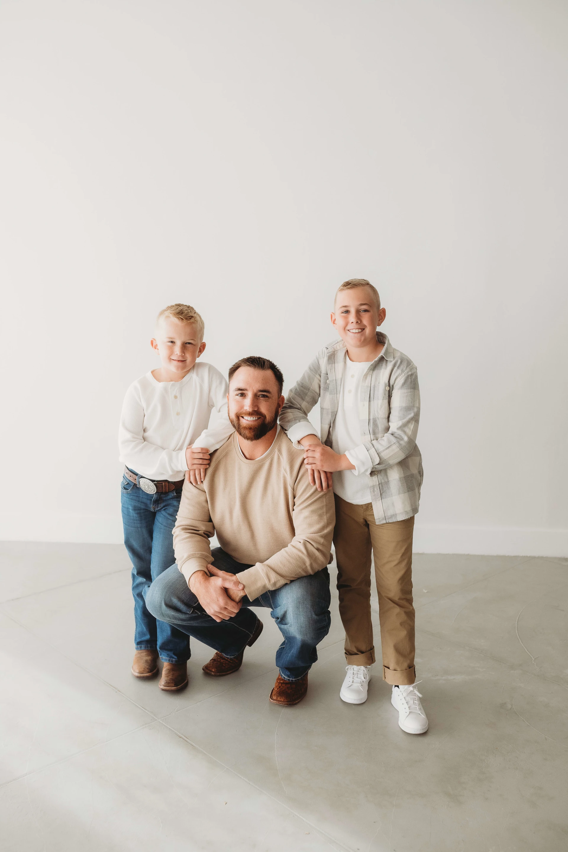 A man squatting on the floor with two boys standing beside him, one on each side, inside a bright, minimalistic room. The man and boys are wearing casual attire, with smiles on their faces.