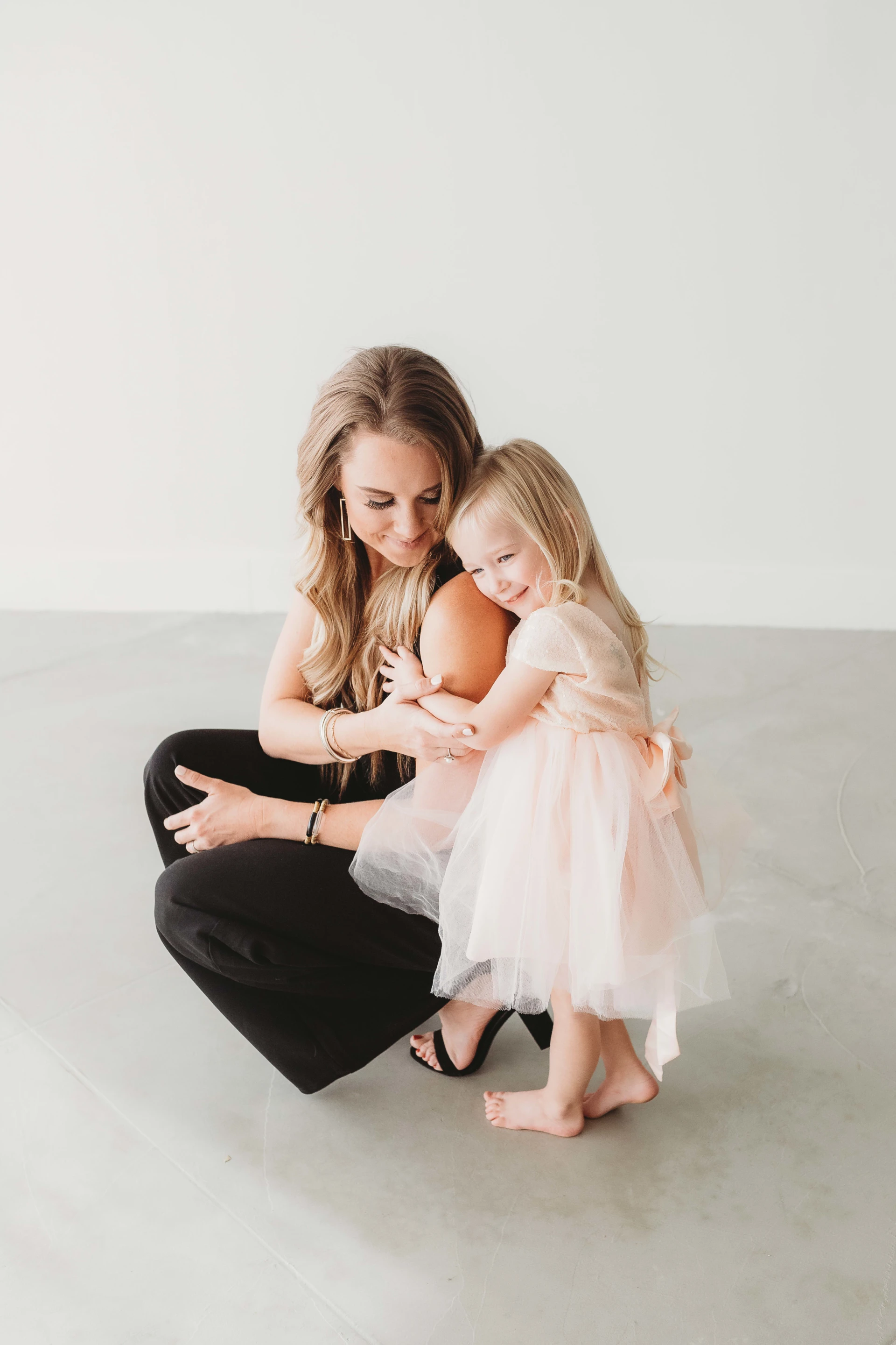 A woman crouching on the floor lovingly embraces a smiling little girl who is wearing a pink dress.