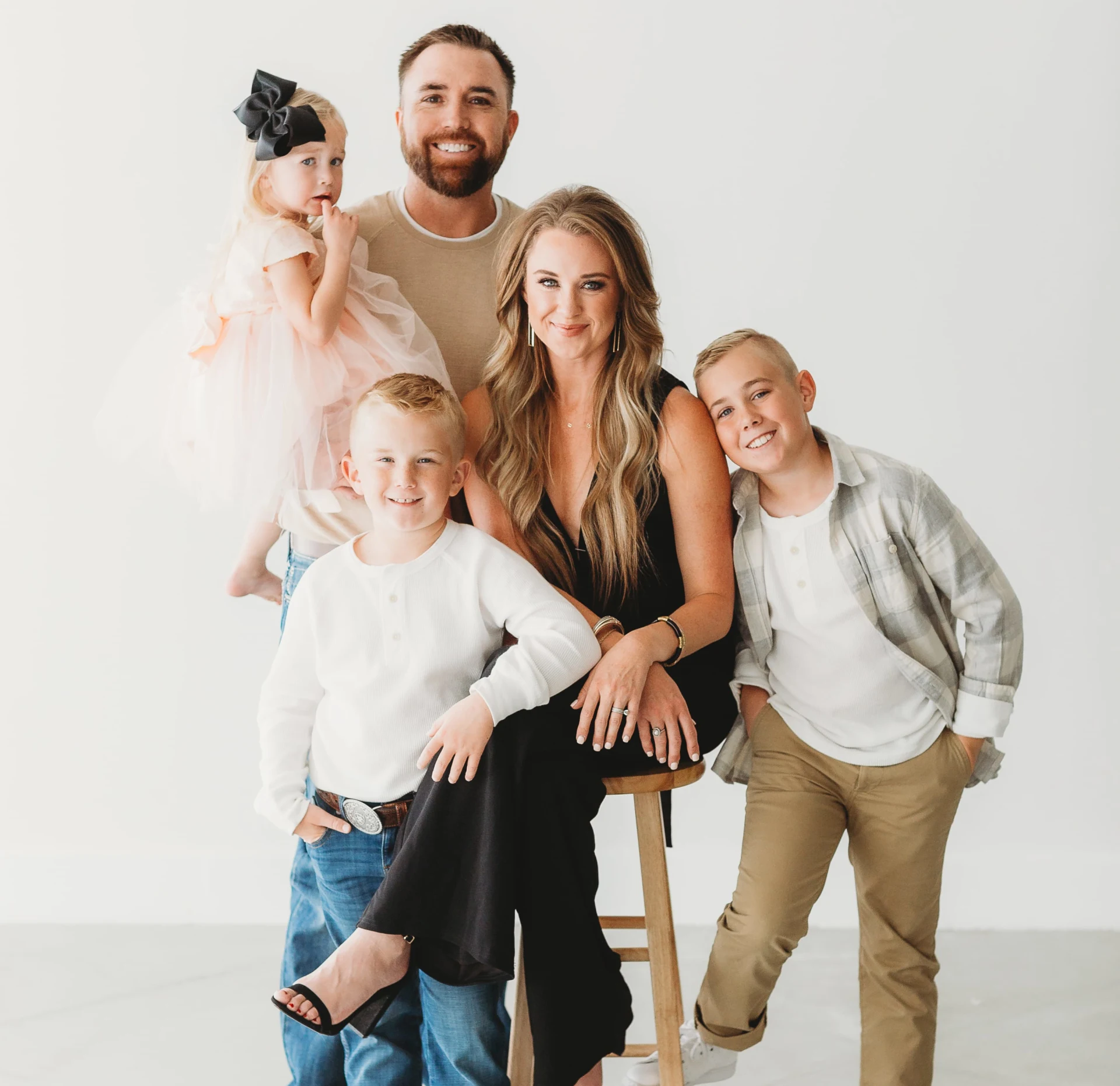 Family portrait with a man holding a young girl, a woman sitting on a stool, and two young boys standing next to her, all smiling in front of a white background.