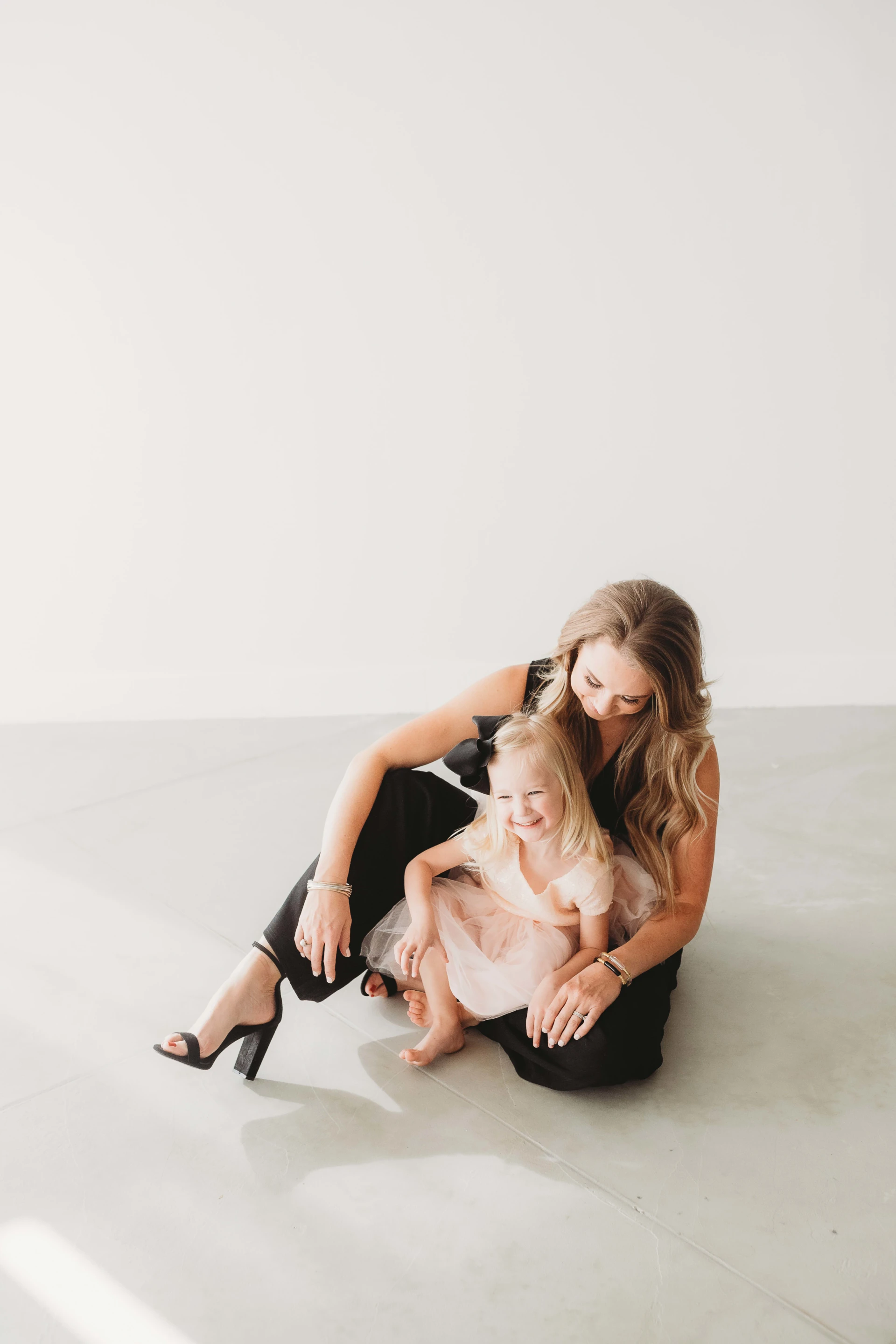 Woman in black outfit sitting on the floor with a smiling young girl in a pink dress in her lap against a white background.