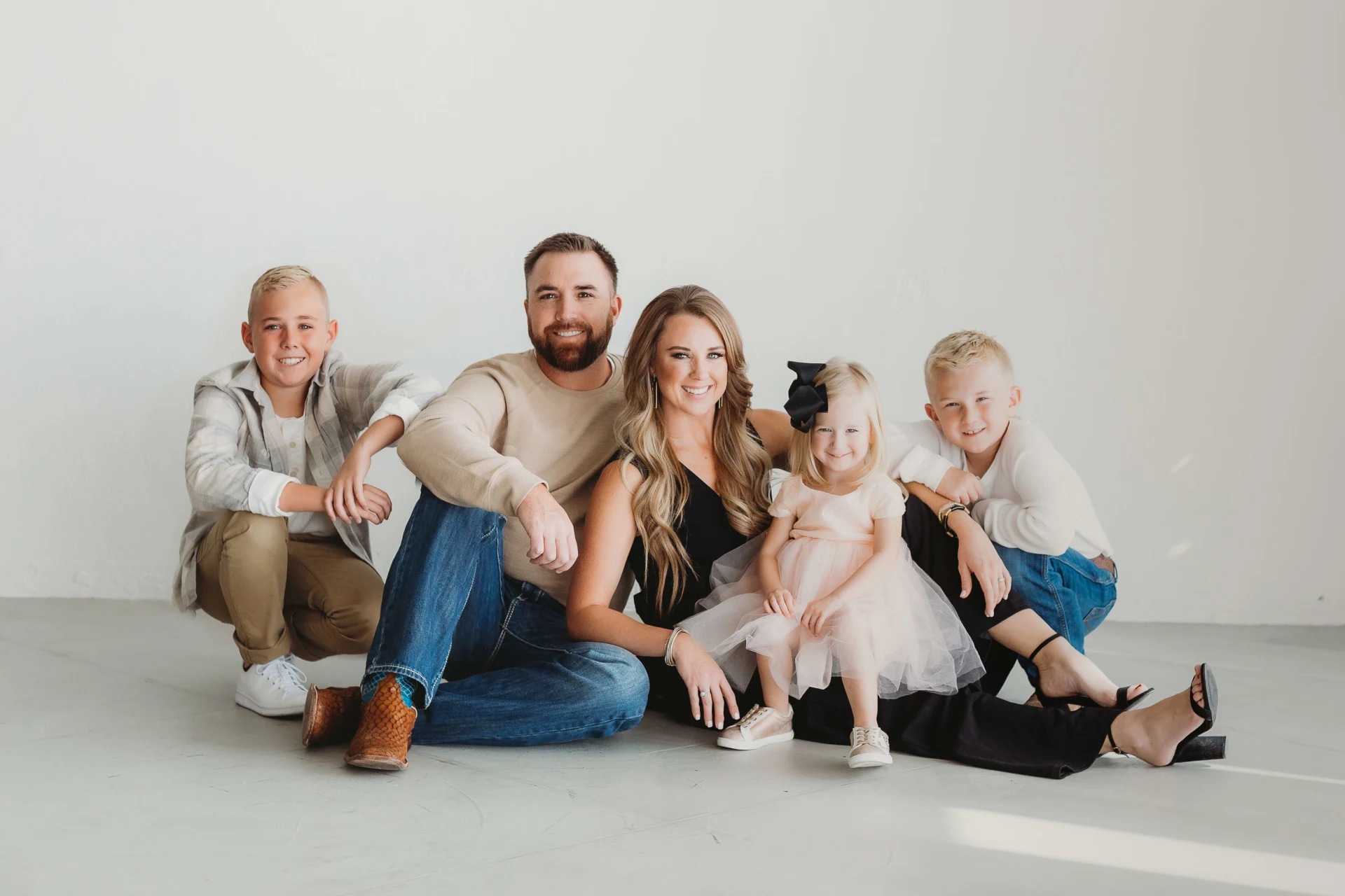 A smiling family of five sitting on the floor against a white background. The man has a beard and is wearing a beige sweater and jeans, the woman has long hair and is in a black dress. There are three children, two boys dressed casually and a young g