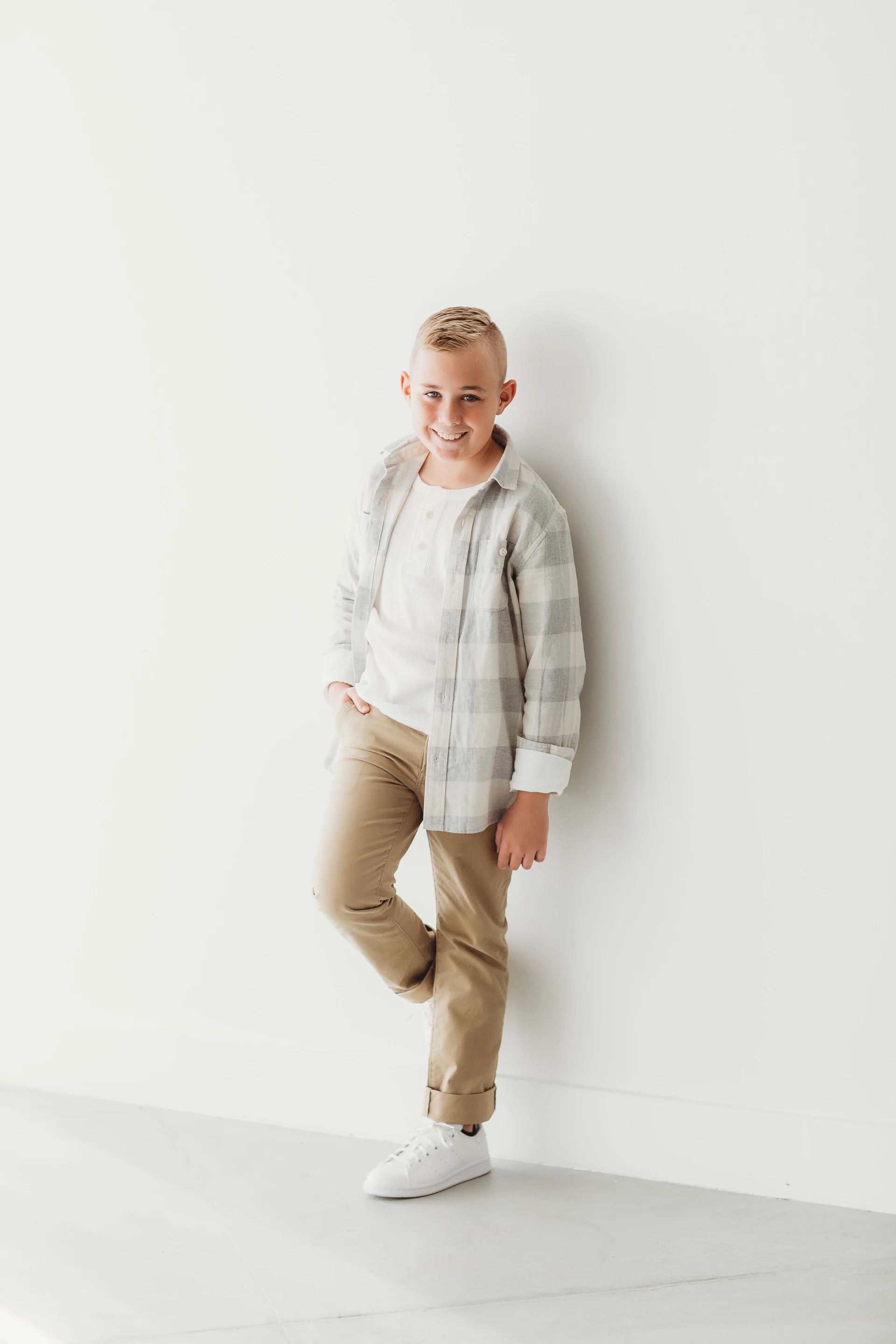 A young boy standing against a white wall, wearing a plaid shirt, white t-shirt, khaki pants, and white sneakers, smiling and posing confidently.