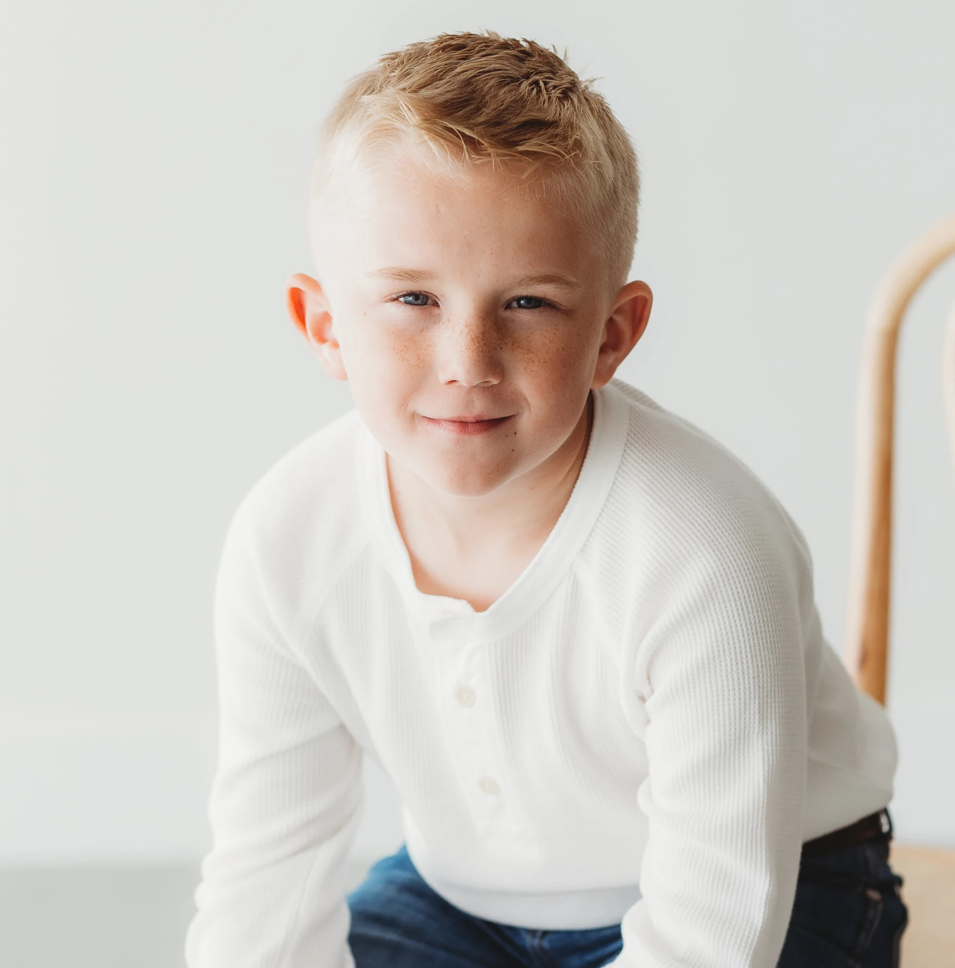Young boy with short blonde hair wearing a white shirt and jeans, sitting on a wooden chair.