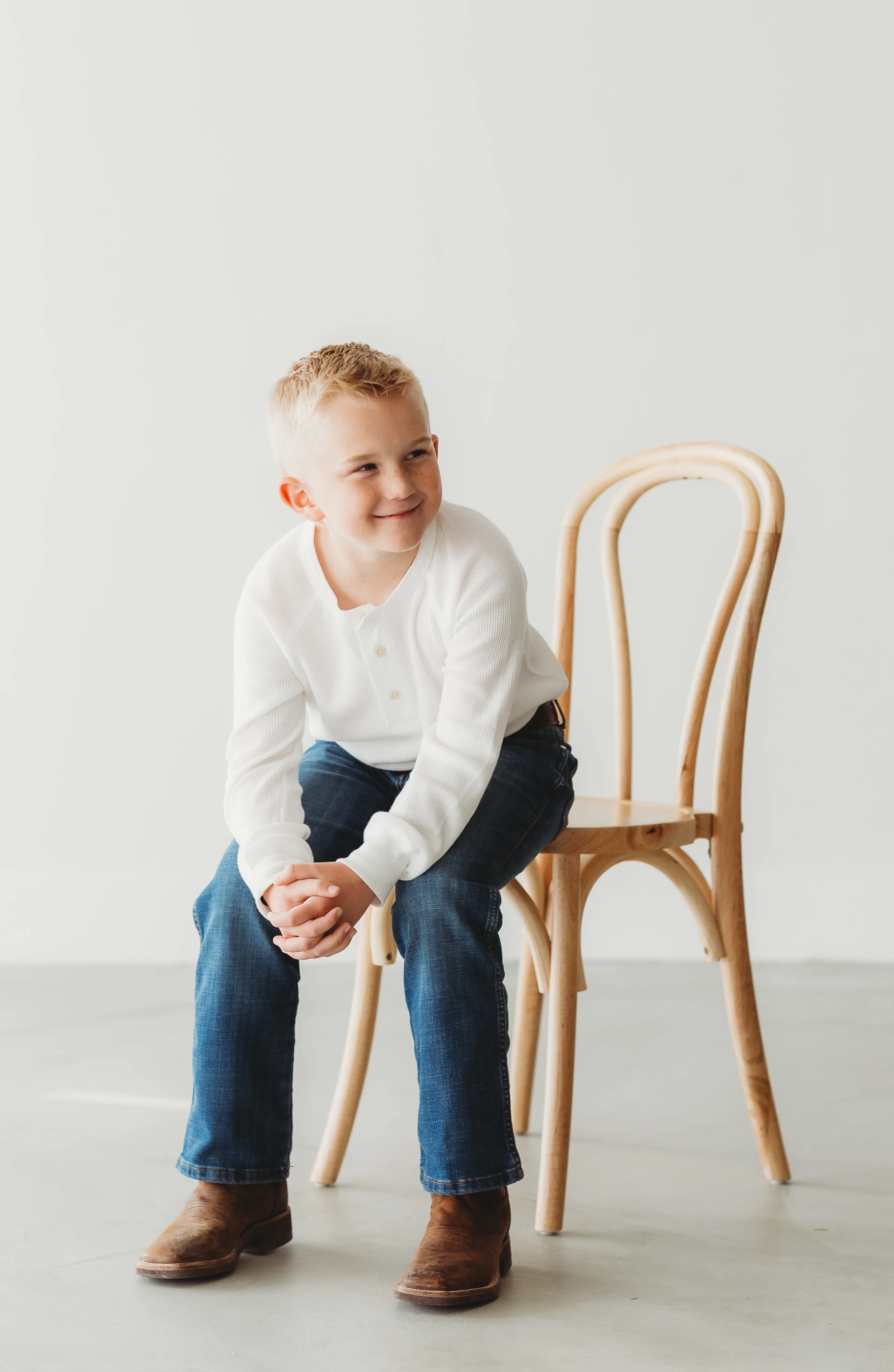 Smiling young boy sitting on a wooden chair, wearing a white long-sleeve shirt, jeans, and brown boots.