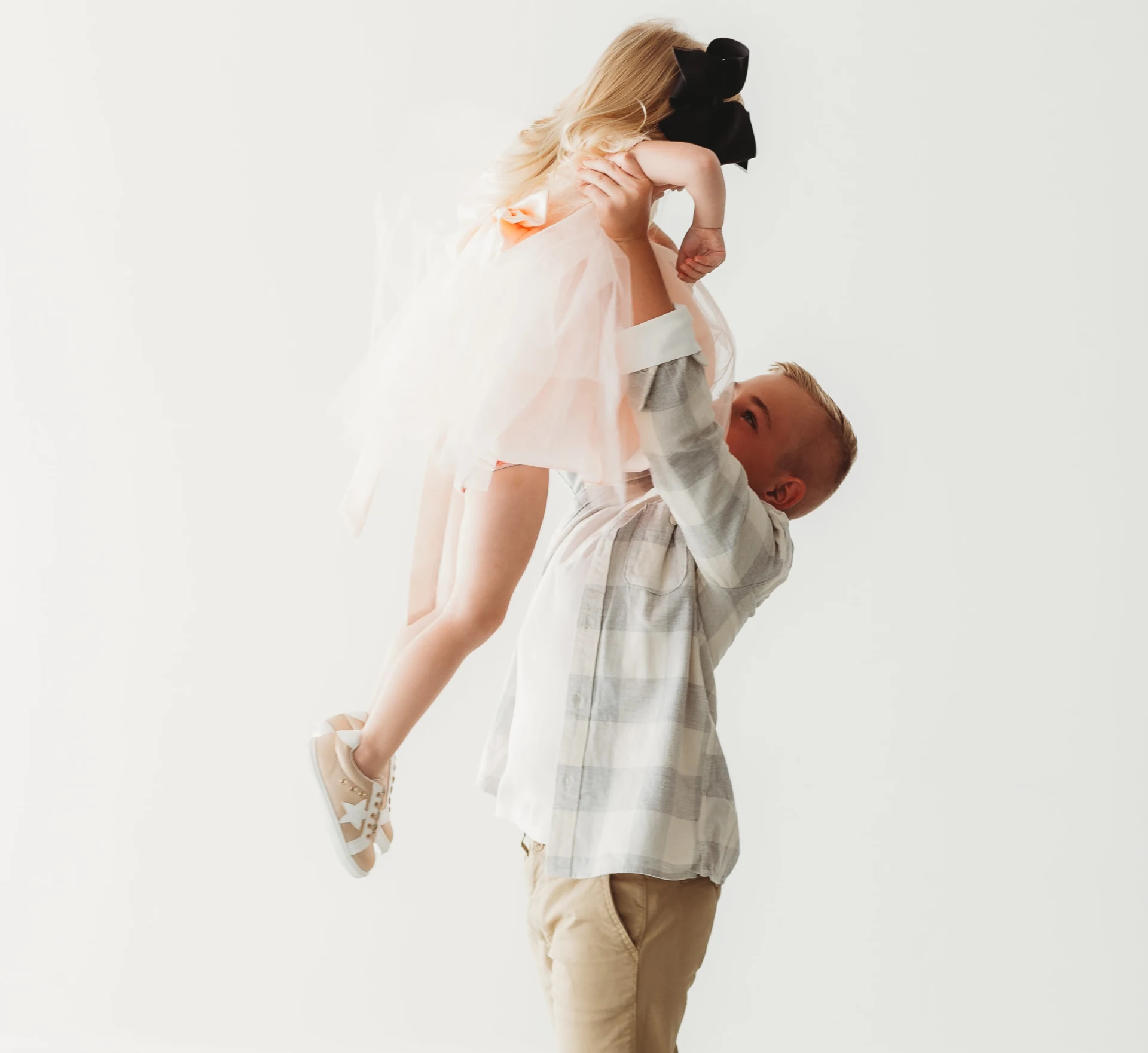 A man lifting a young girl wearing a pink dress and black bow, with a light background.