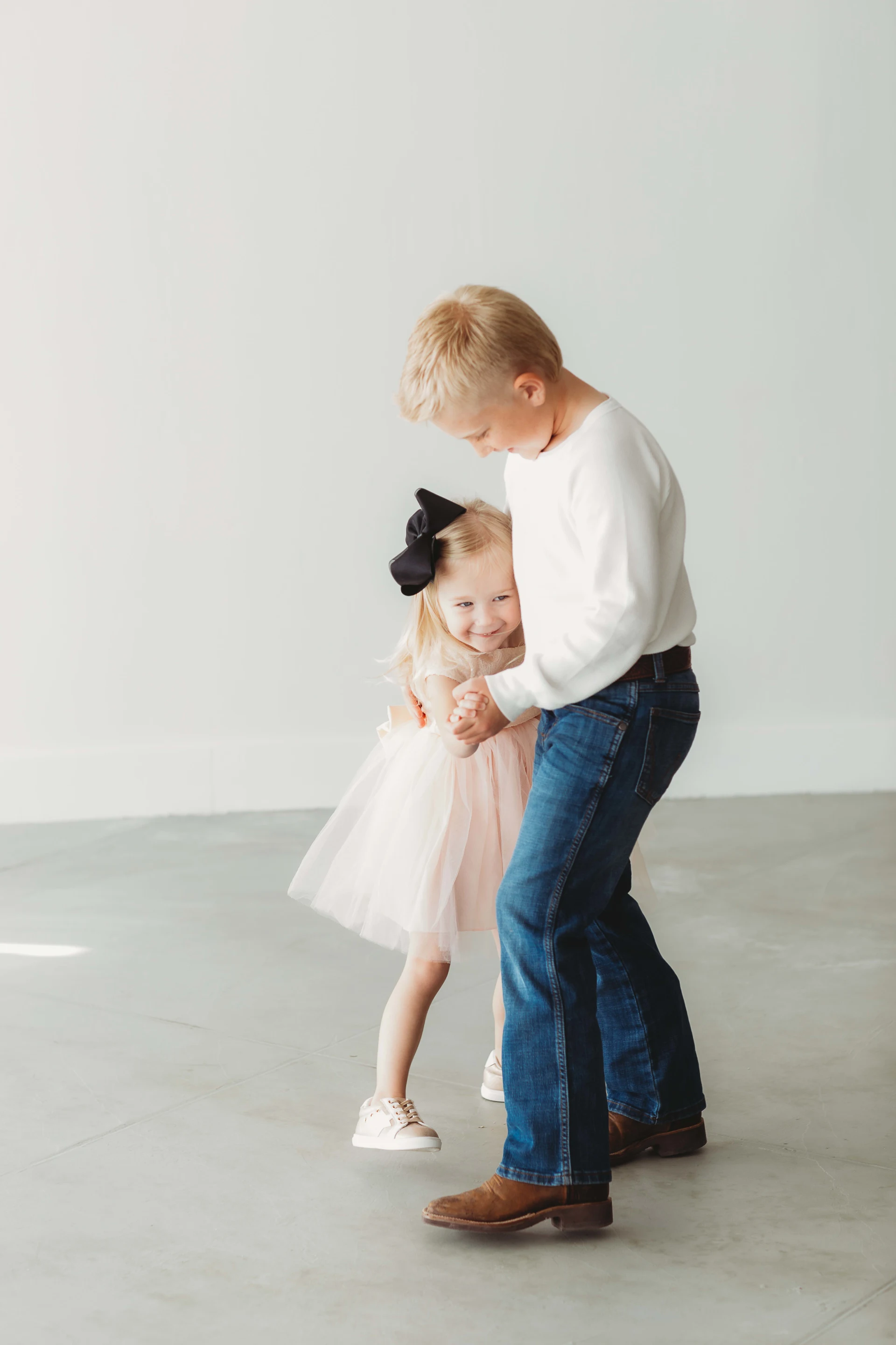 A young boy hugging a little girl, both smiling, in a bright room. The girl wears a pink dress and black hair bow, the boy wears a white shirt and jeans.