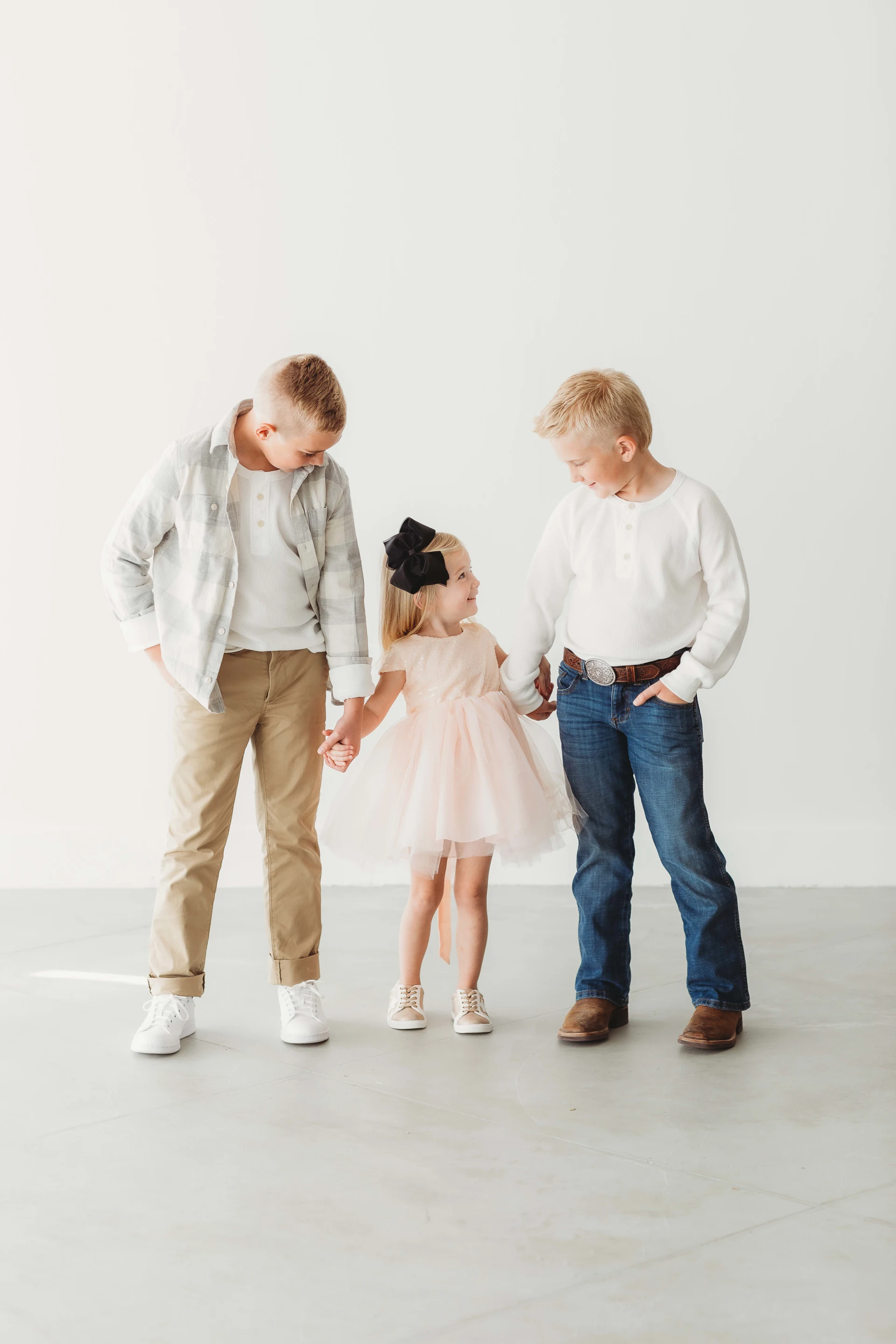 Three children standing and holding hands, with two boys on either side of a girl in a pink dress with a black bow. One boy wears a plaid shirt and khaki pants, the other wears a white long-sleeve shirt and jeans.