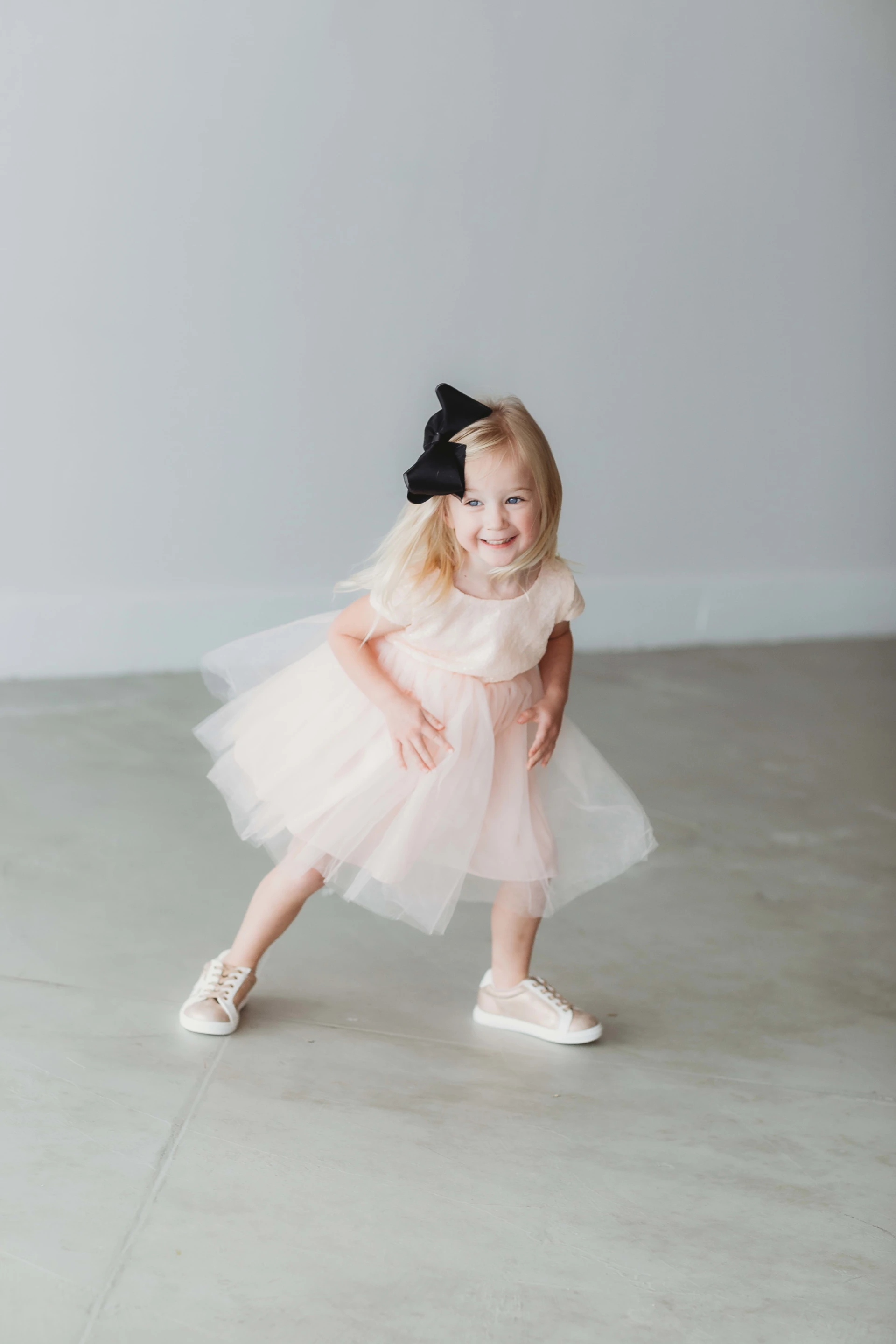 Young child smiling, wearing a light pink dress and a black bow, standing on a gray floor.