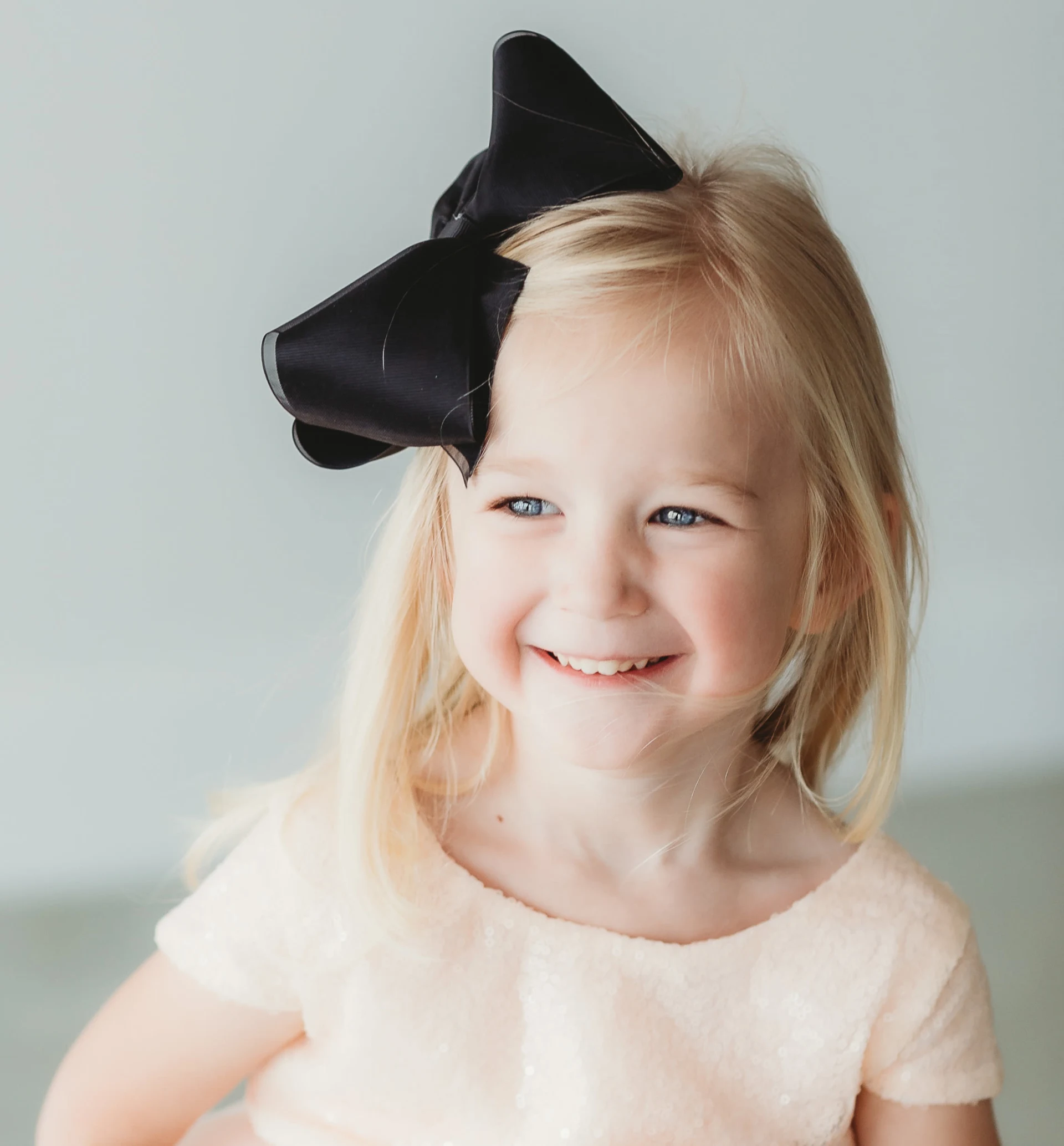 Smiling blonde girl with a black bow in her hair wearing a light peach dress.