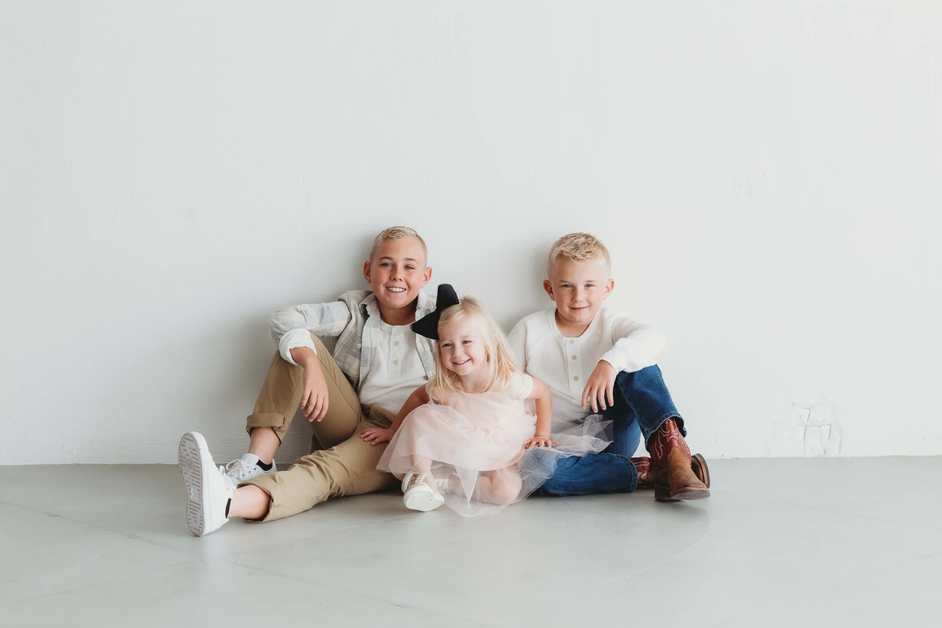 Three children sitting on the floor against a white wall, smiling. Two boys are wearing casual clothes, and a girl is in a pink dress with a black bow in her hair.