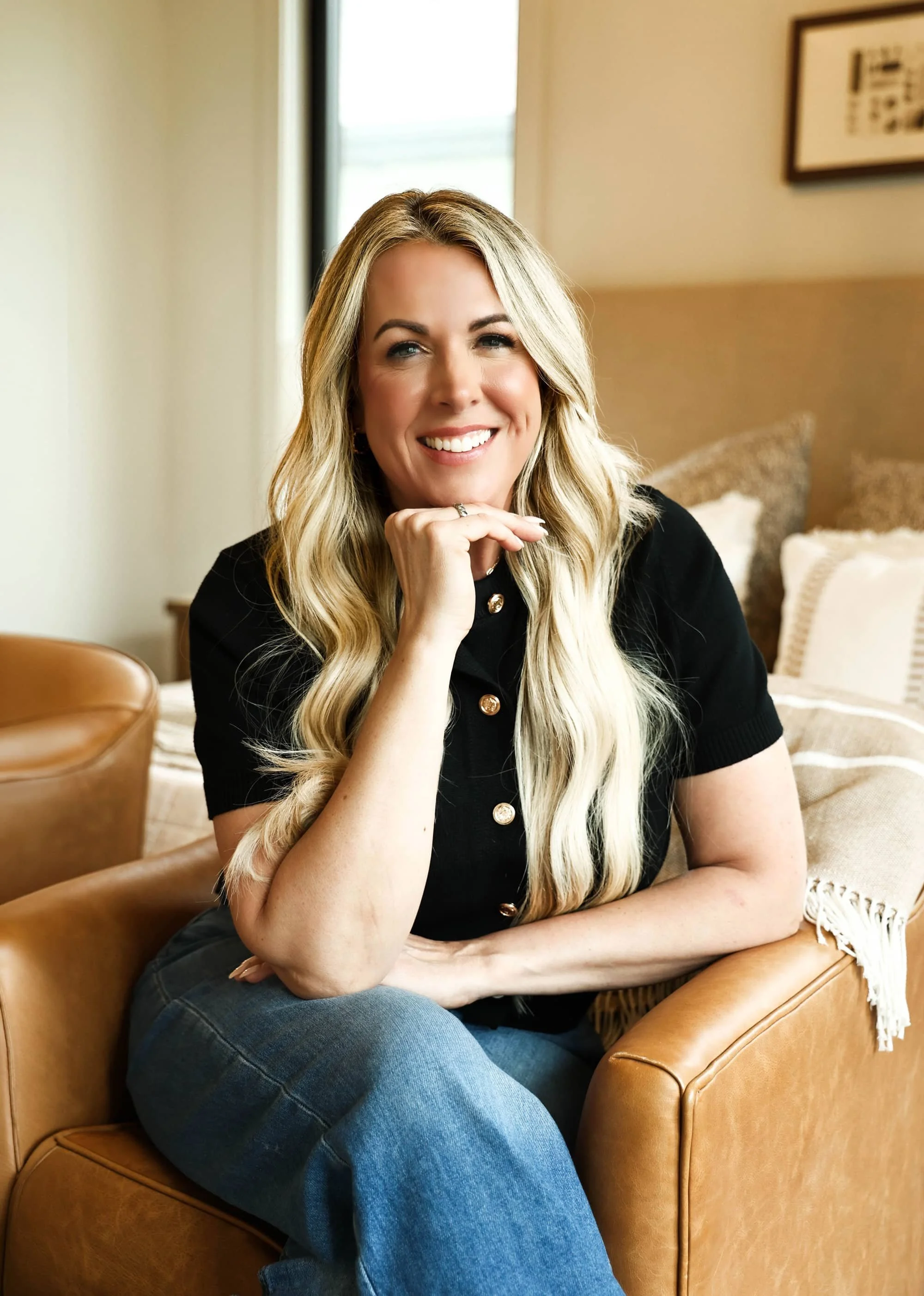 A woman with long blonde hair, wearing a black shirt with gold buttons and blue jeans, smiling and resting her chin on her hand, sitting on a tan leather armchair in a cozy room with beige walls, pillows, and framed artwork in the background.
