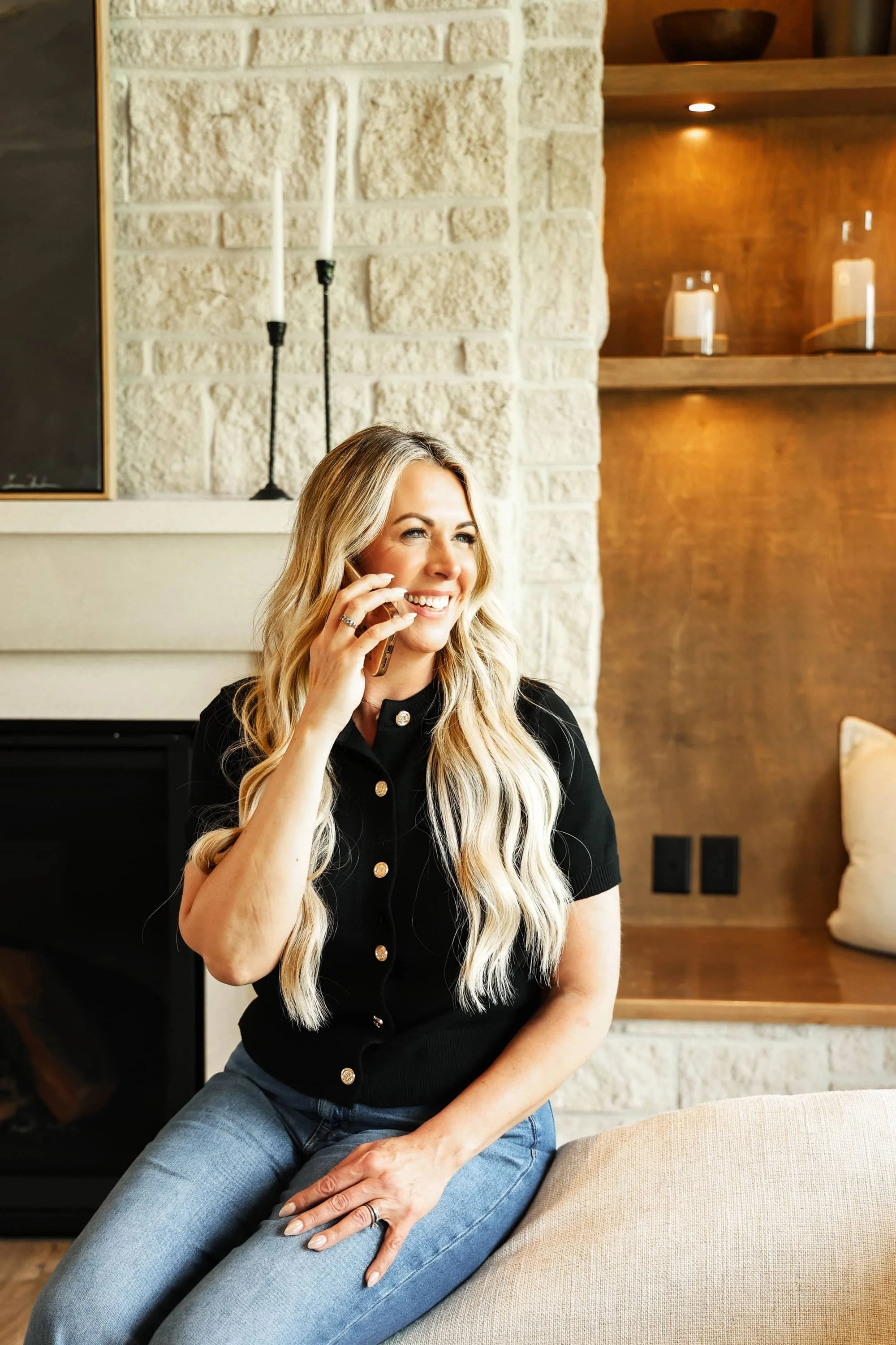 A woman with long blonde hair, wearing a black shirt with gold buttons, sitting on a beige couch in a cozy living room and talking on a mobile phone.