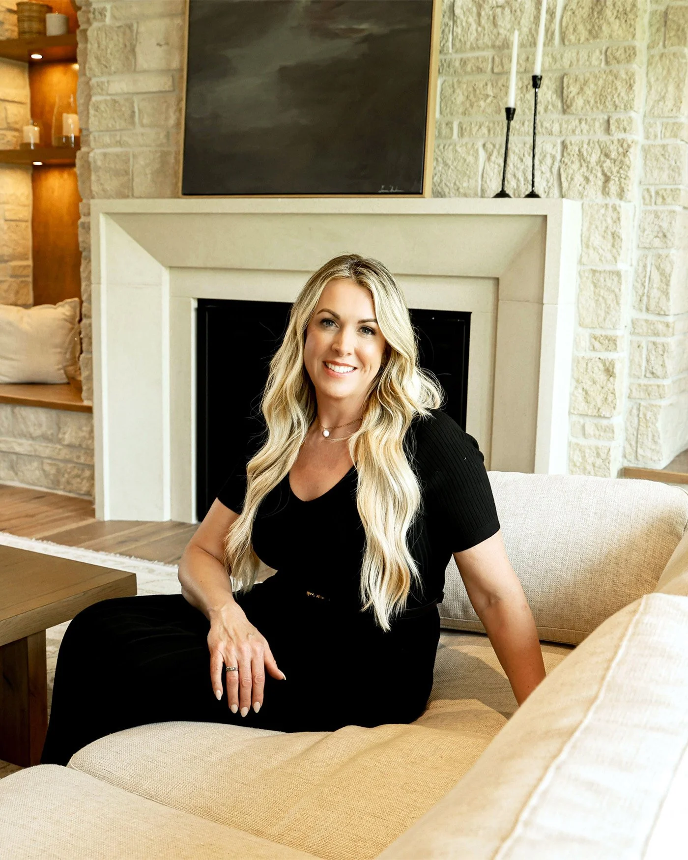A woman with long blonde hair, wearing a black outfit, sitting on a beige couch in a living room with a stone fireplace and a blank blackboard above it.