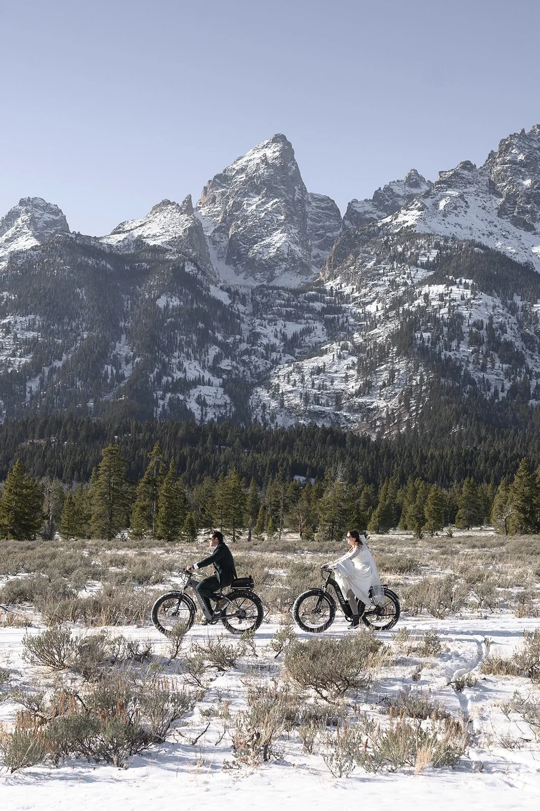 a couple riding bikes in their wedding outfits though the snow in the Grand Tetons