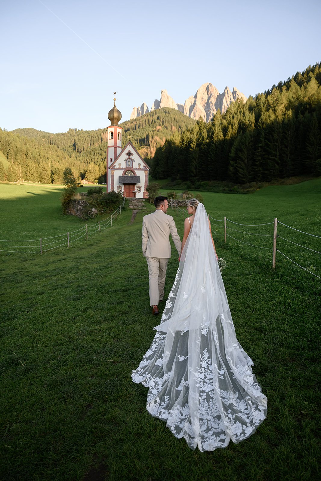 a couple walking through a field in the italian dolomites with a church behind them on their wedding day