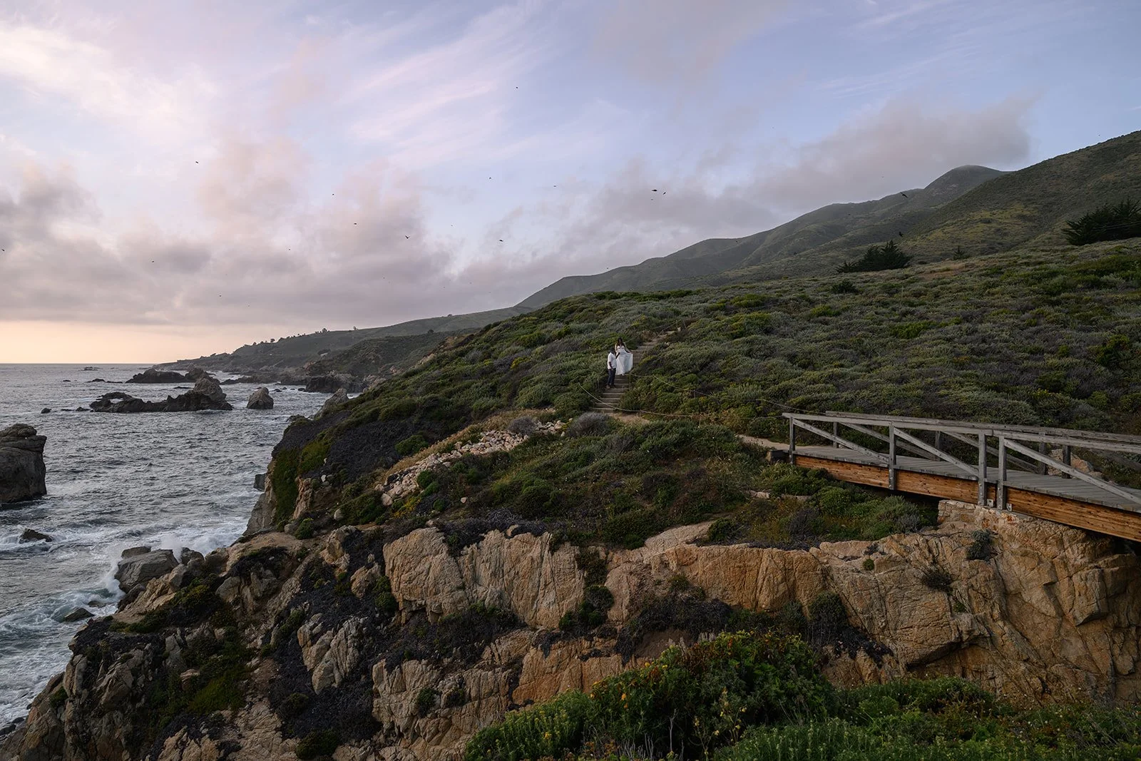 sunset in big sur with a couple walking down the steps in the hill next to a bridge with the coast line stretching out and birds flying around them at sunset