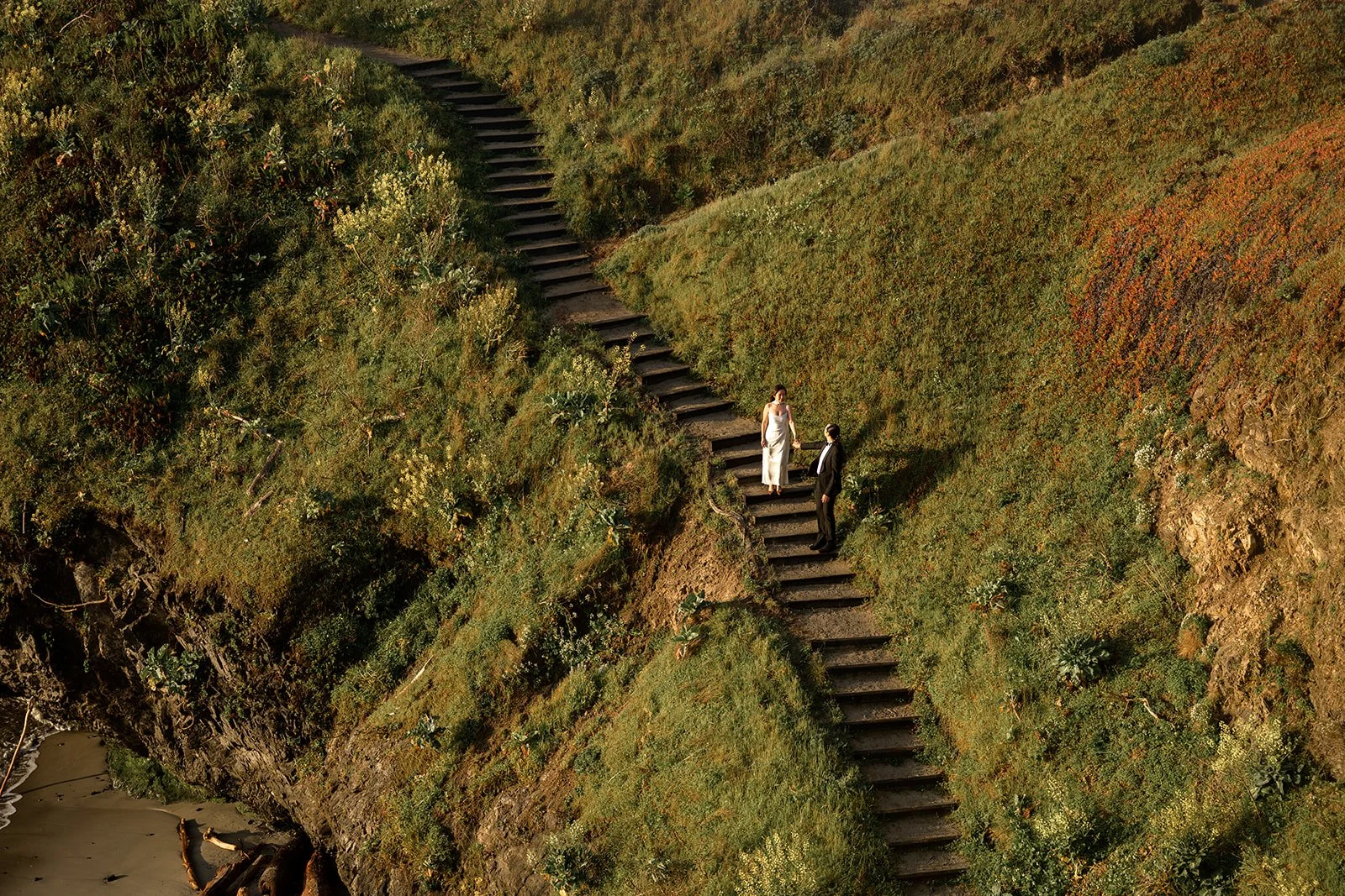 a photo of a bride and groom from above holding hands and walking down a long staircase on the side of a green hill in mendocino