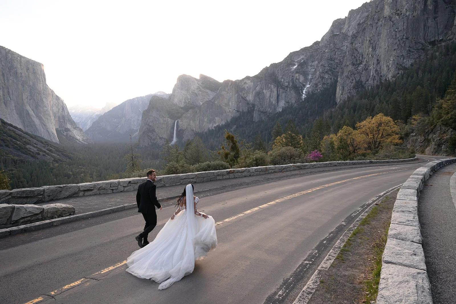 a photo of a bride and groom running down a street in yosemite national park with yosemite valley behind them at sunrise